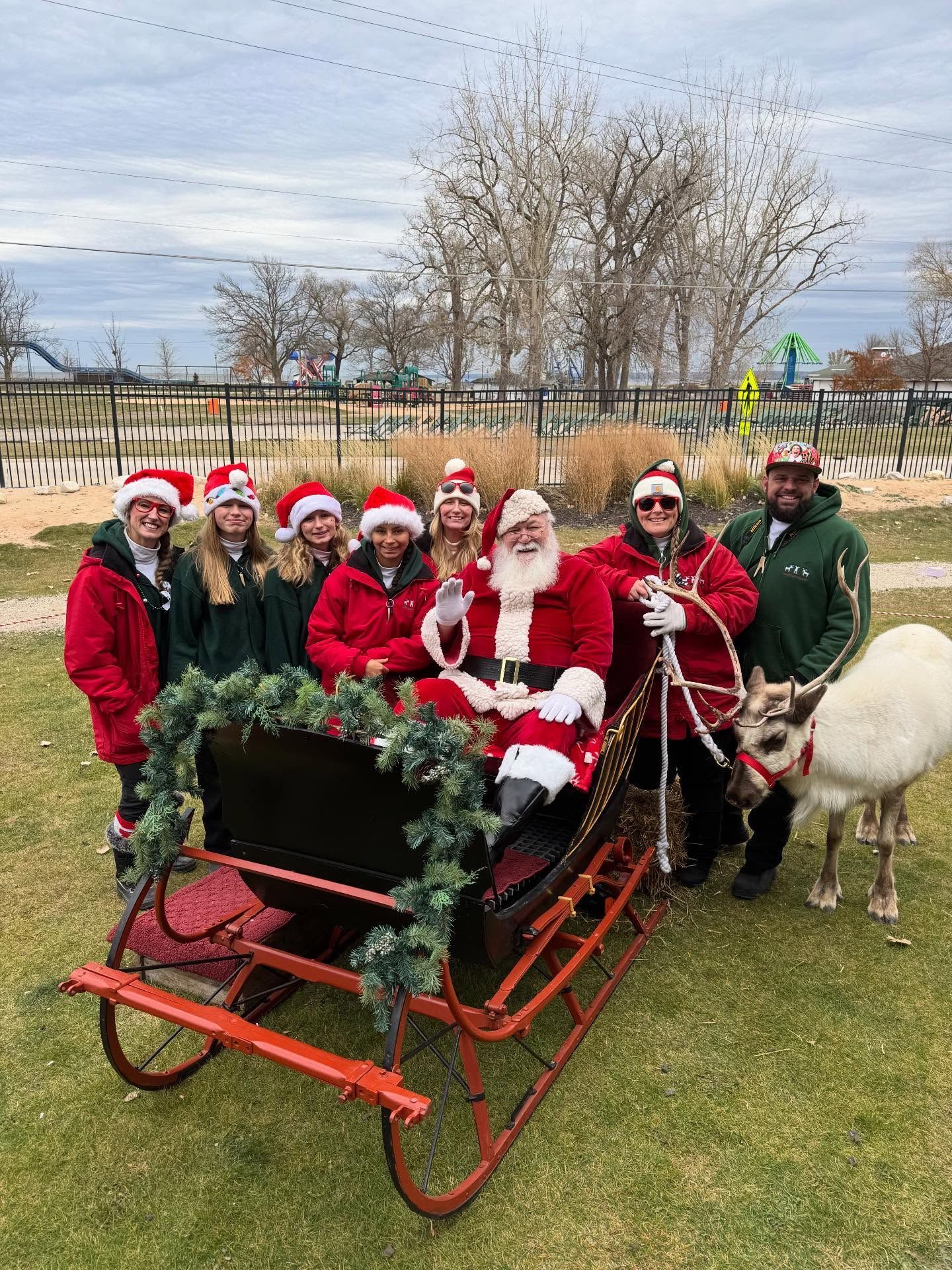 Santa sits in a red sleigh with elves outdoors; a reindeer stands nearby. Everyone smiles.