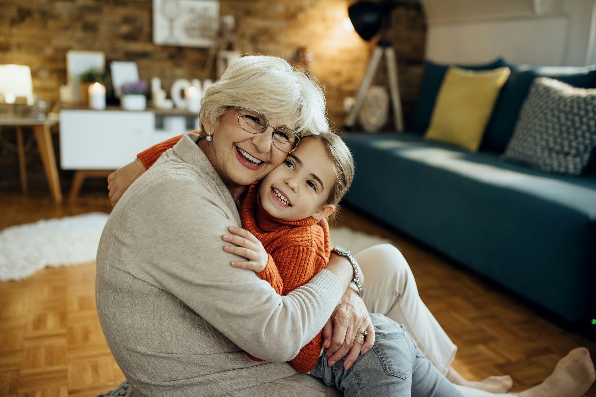 Two people hugging on a living room floor, smiling warmly with a couch and decor in the background.