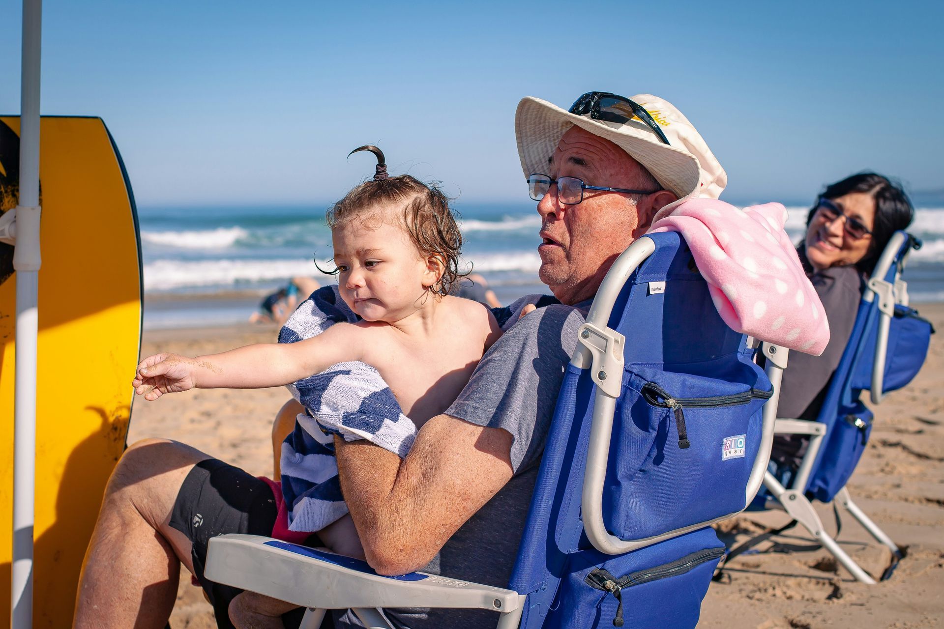 Two people sit in beach chairs, one holding a child on a sunny sandy beach.