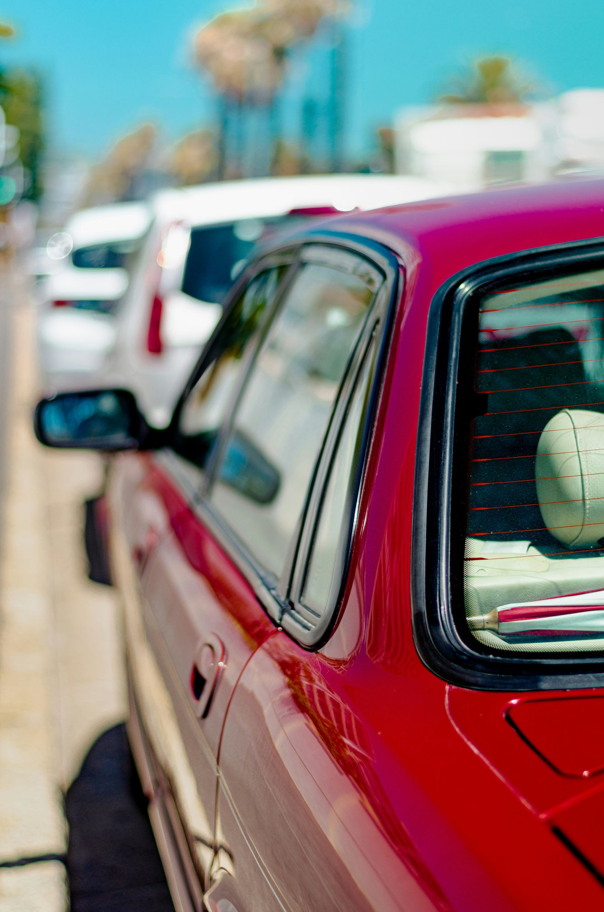 Red car parked along a sunny street, with a white car ahead and blue sky in the background