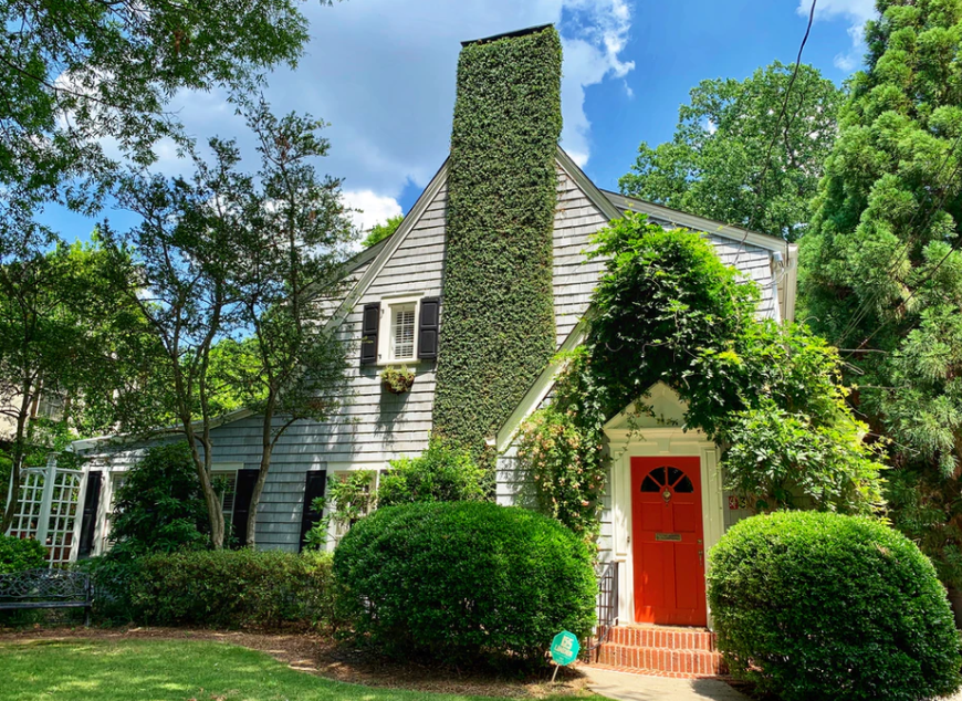 Cottage with gray shingle siding and a tall, ivy-covered chimney. Red front door, surrounded by greenery, under blue sky.