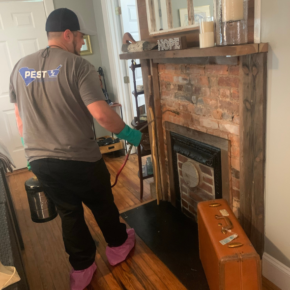 Pest control worker sprays fireplace. Brown brick, wooden mantel. He wears a hat, gloves, booties.