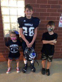 Three kids in front of a brick wall: a football player, a girl, and a boy, all wearing masks.