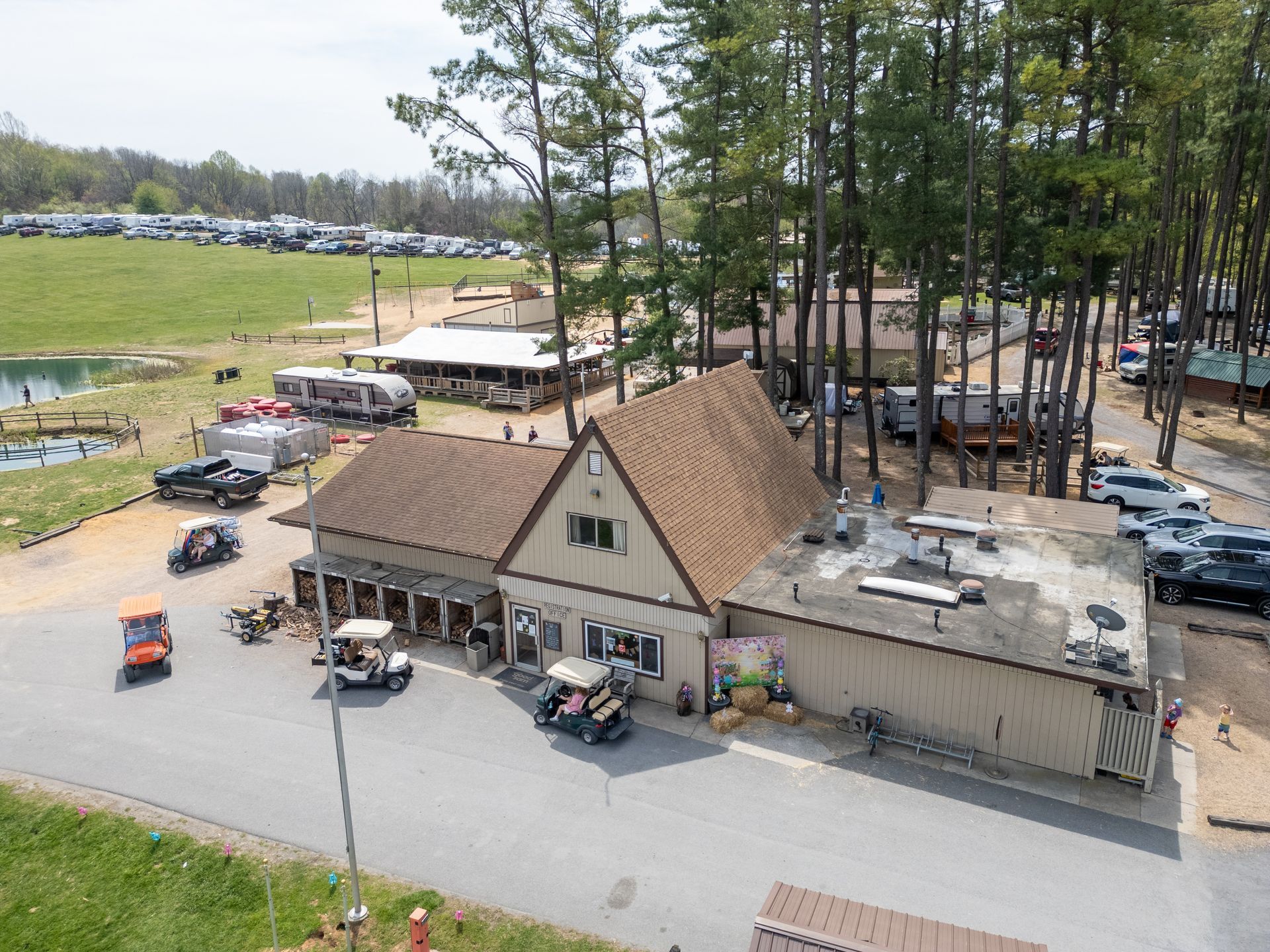 An aerial view of a campground store with a brown-roofed building, golf carts, and parked vehicles near a grassy field.