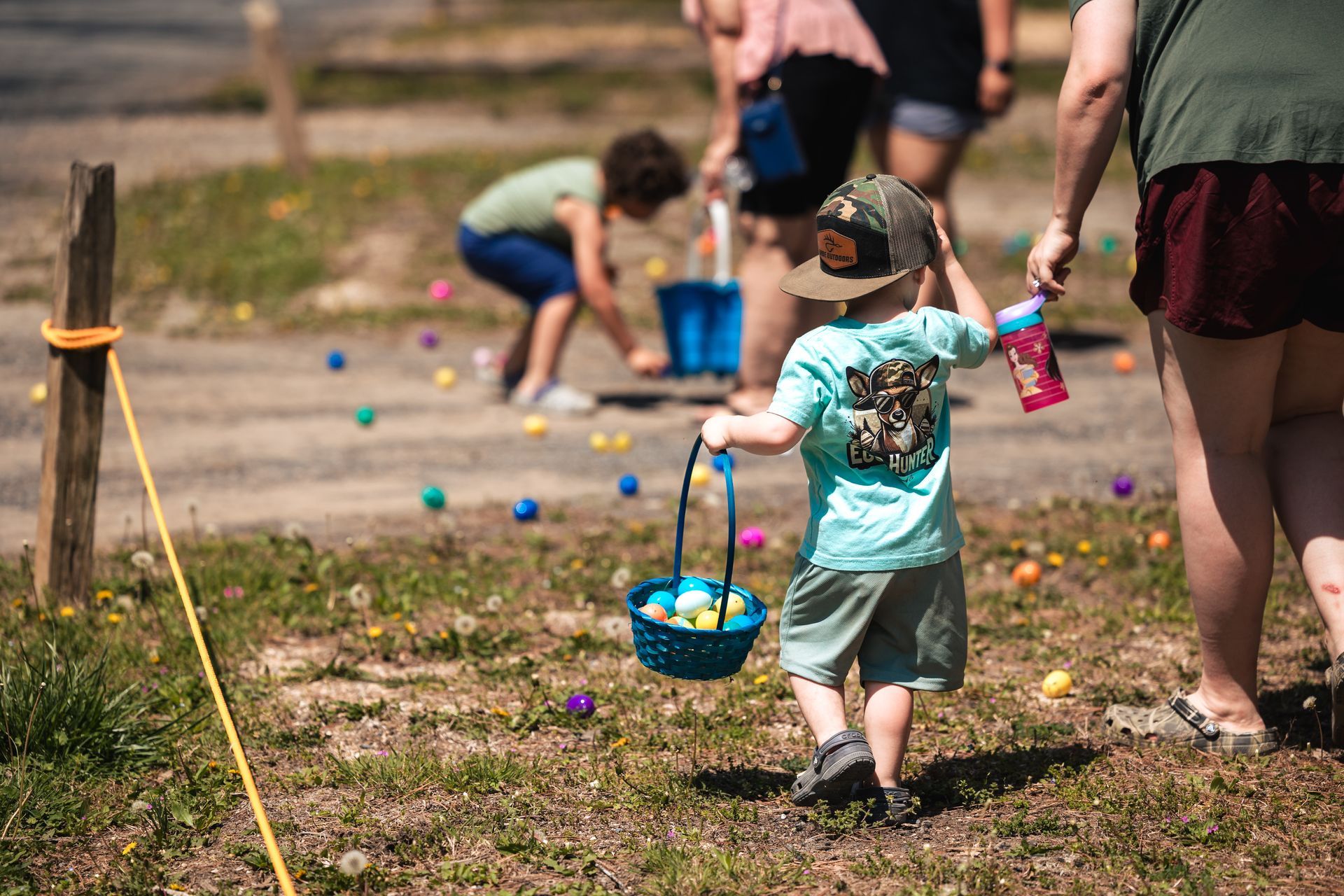 Child in teal shirt carries basket, walking through field, Easter eggs scattered.