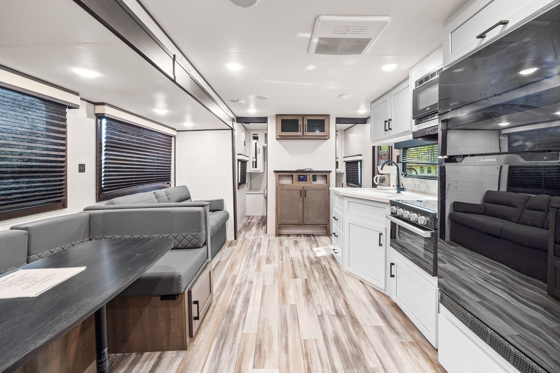Interior of a modern recreational vehicle showing a grey dining booth, kitchen area with an oven, and a black sofa.