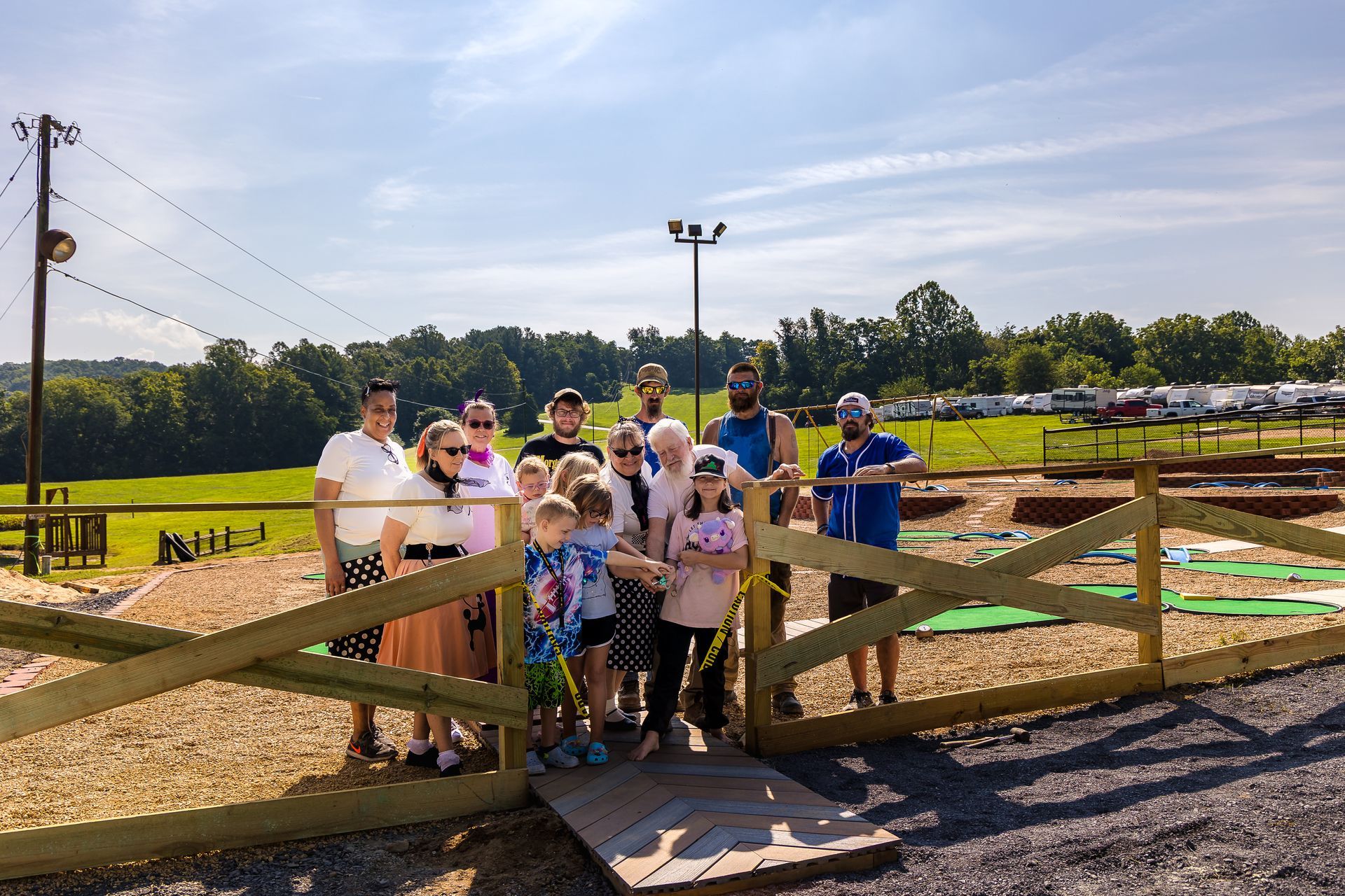 A group of people gathers behind a wooden fence at a miniature golf course on a sunny day.