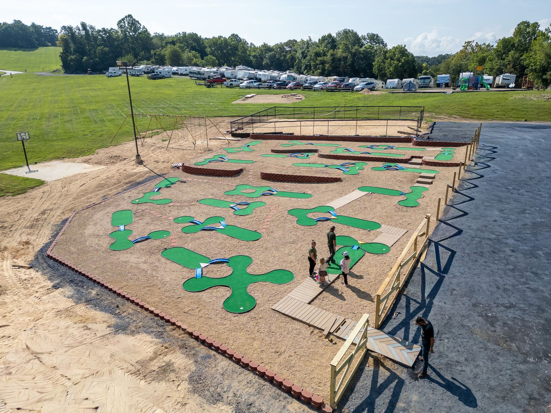 An elevated view of a outdoor mini-golf course with green turfed holes set on a gravel field near trees and parked vehicles.