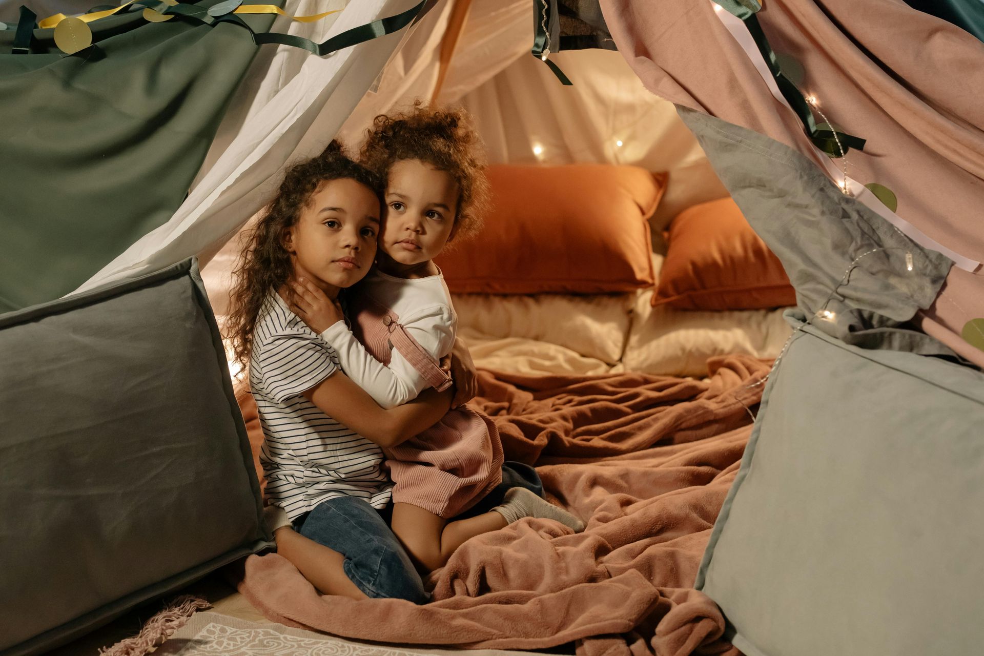 Two children in a cozy fort, hugging, surrounded by pillows and blankets.