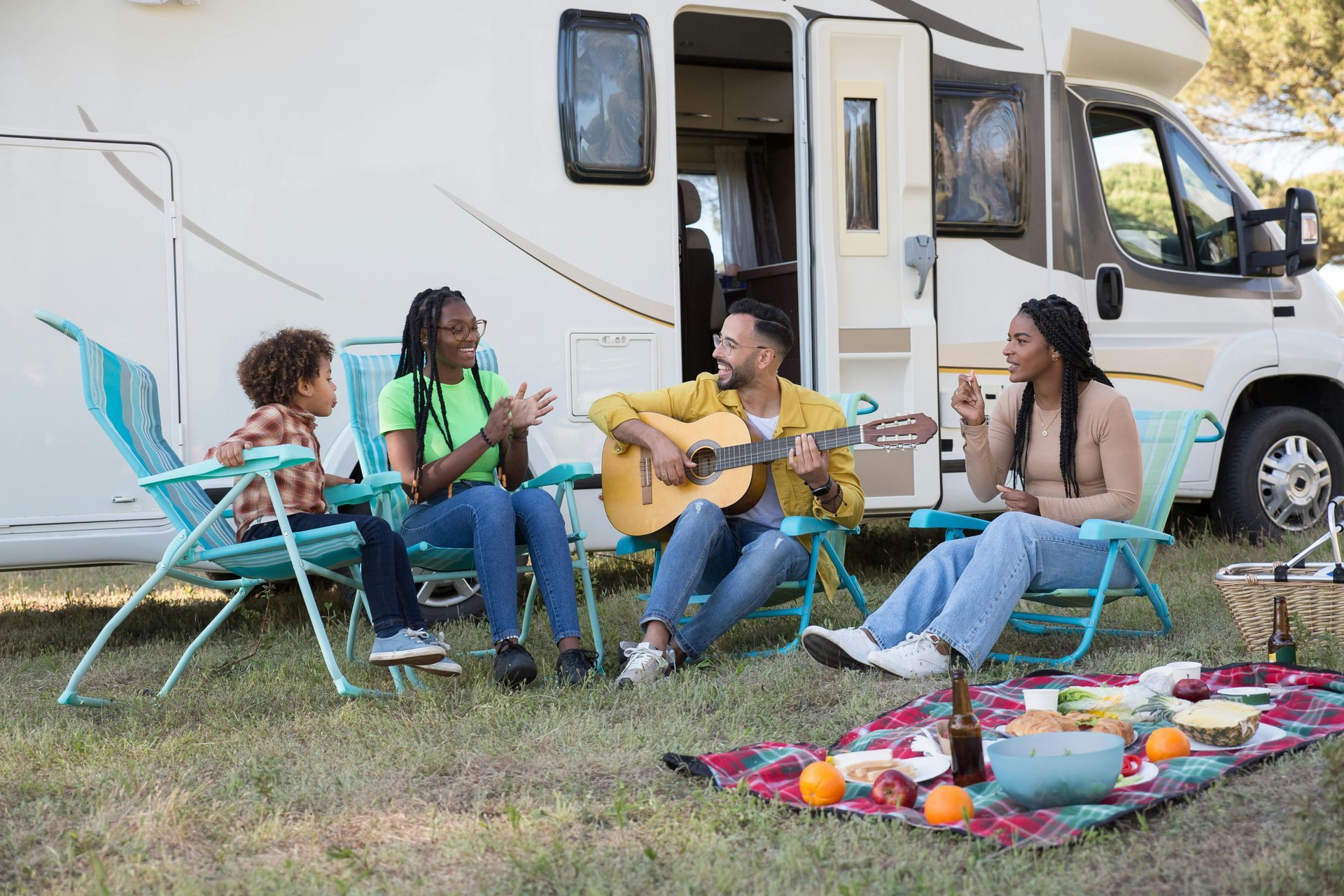 Family camping outside an RV; a person plays acoustic guitar while others sit in lawn chairs near a picnic blanket.