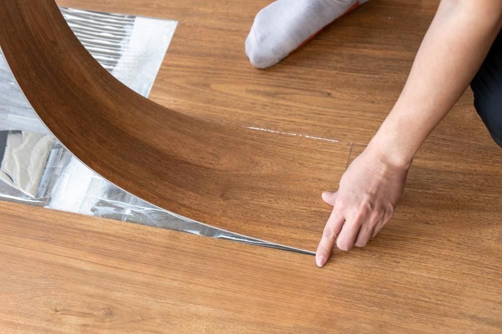 Person installing brown wood-look flooring, pointing to a seam. Gray and white underlayment visible.