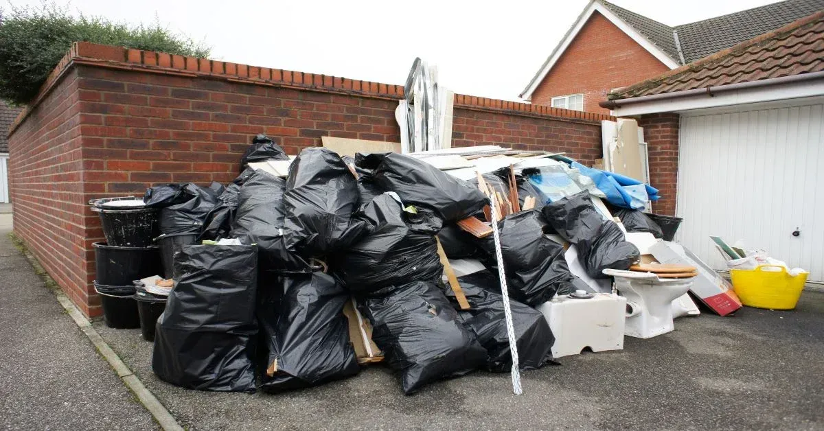 Pile of black trash bags, debris, and a toilet next to a brick wall and garage.