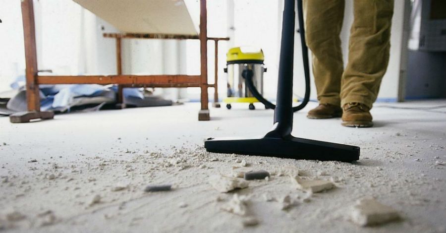 Person vacuuming debris on a concrete floor in a construction setting; yellow vacuum cleaner in background.