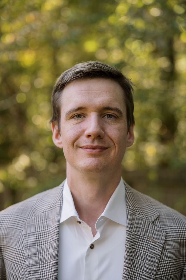 Portrait of a man in a checkered blazer and white shirt, standing outdoors with a blurred autumn background