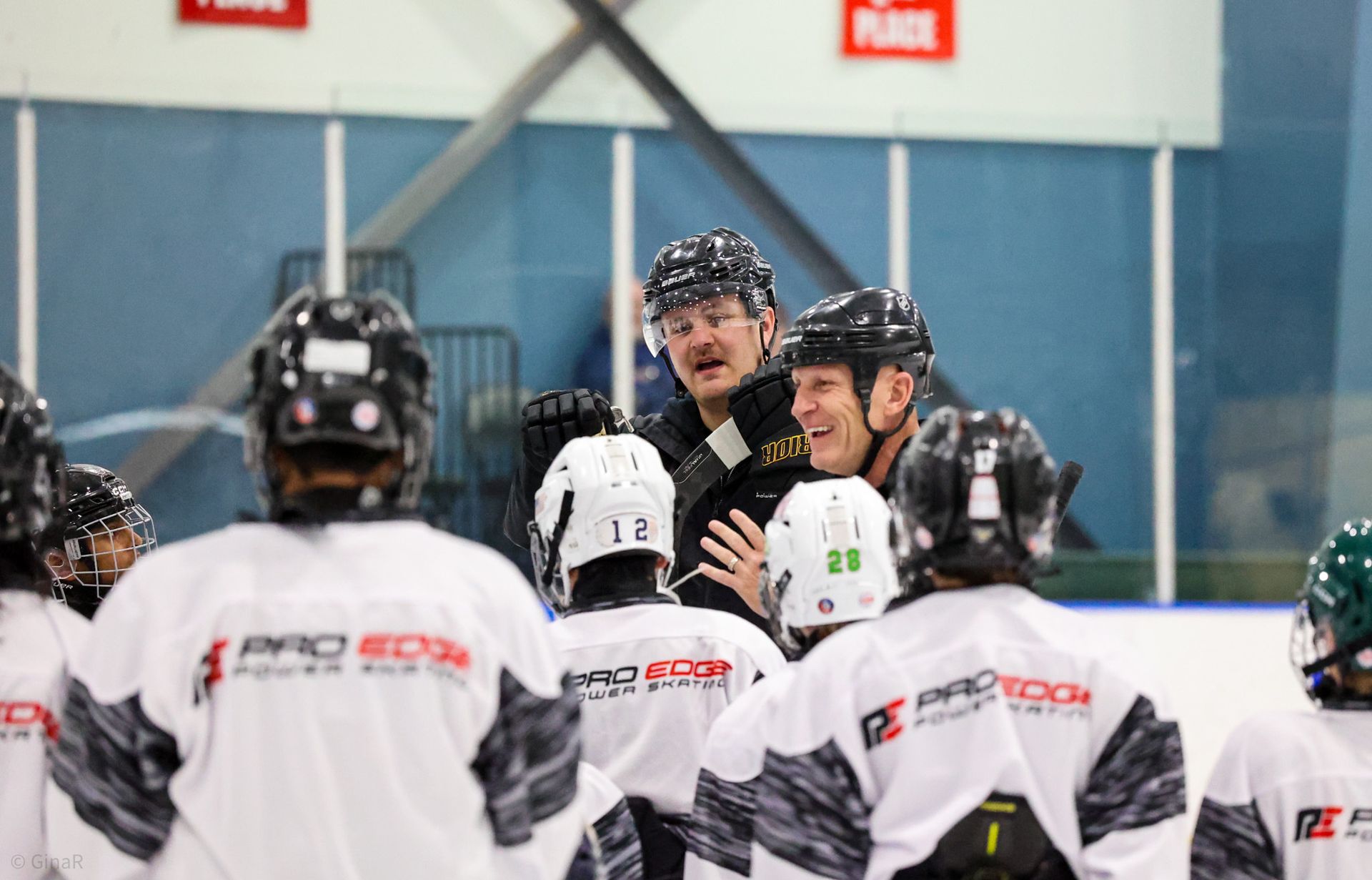 Hockey players in white jerseys gather around a smiling coach on the rink