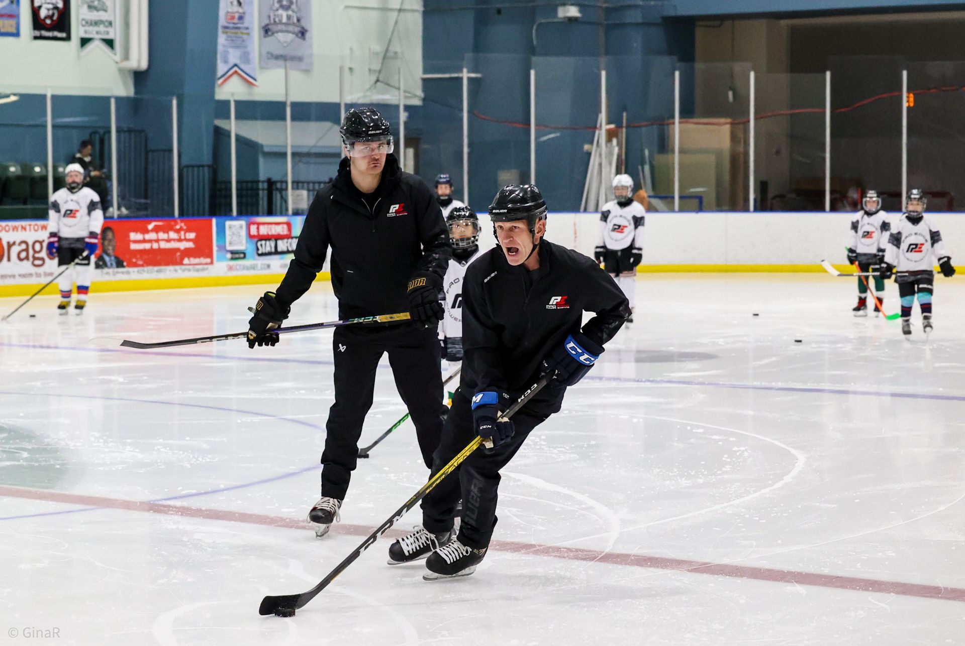 Two hockey players in black practice skating on an indoor rink while others play in the background