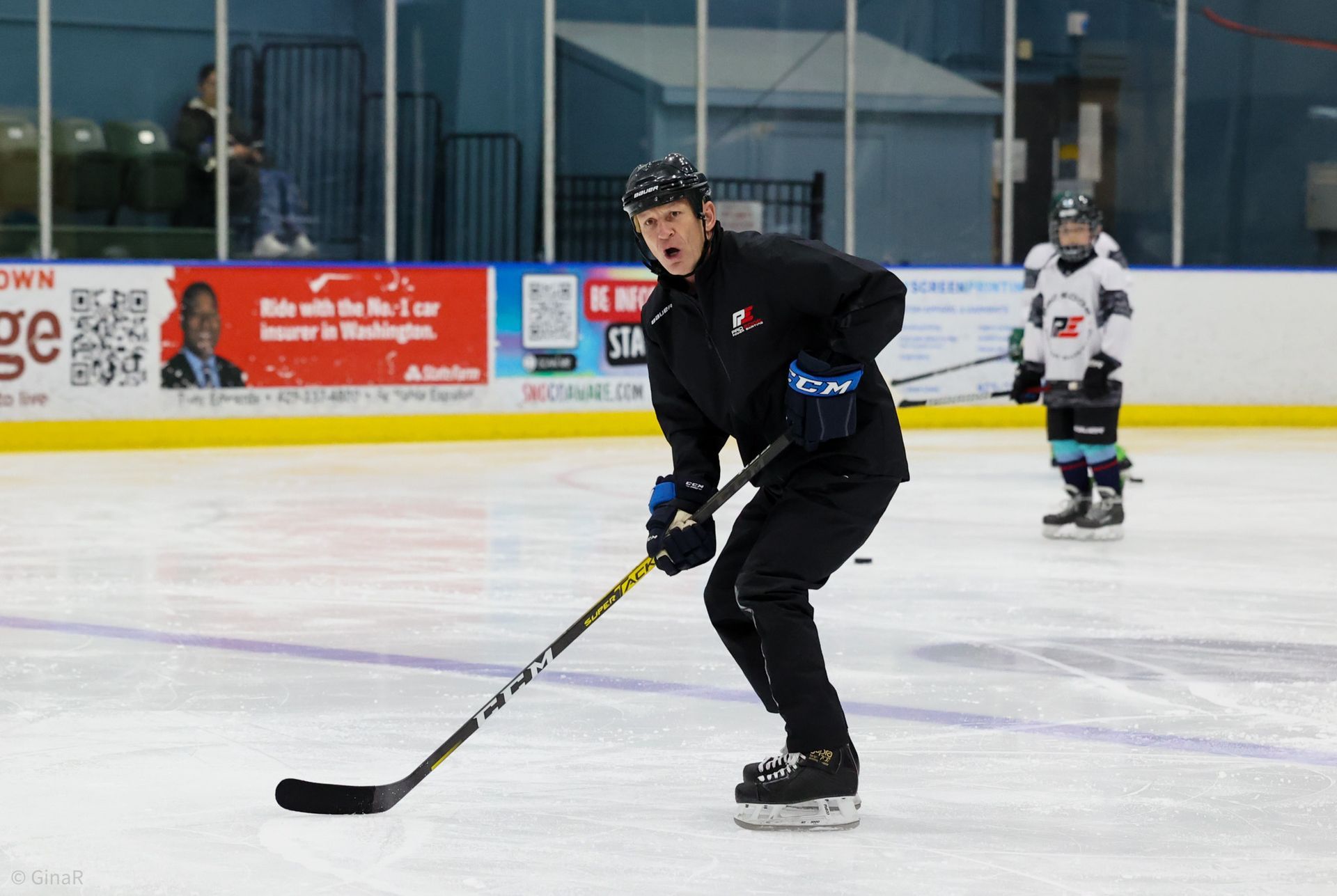 Hockey player in black skating on an indoor ice rink, holding a stick and looking forward.