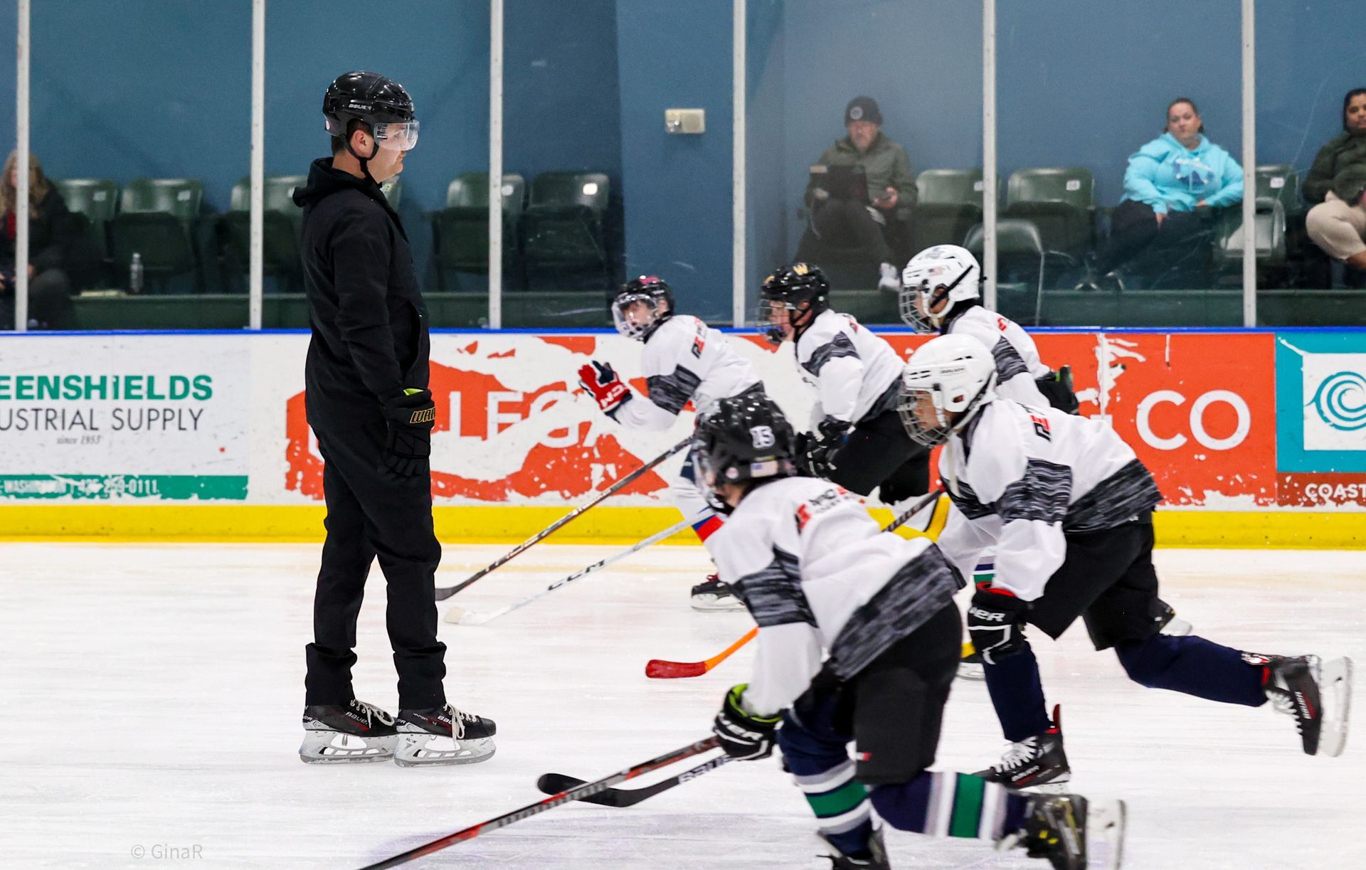 Youth hockey players in white uniforms skate toward a coach on an outdoor ice rink