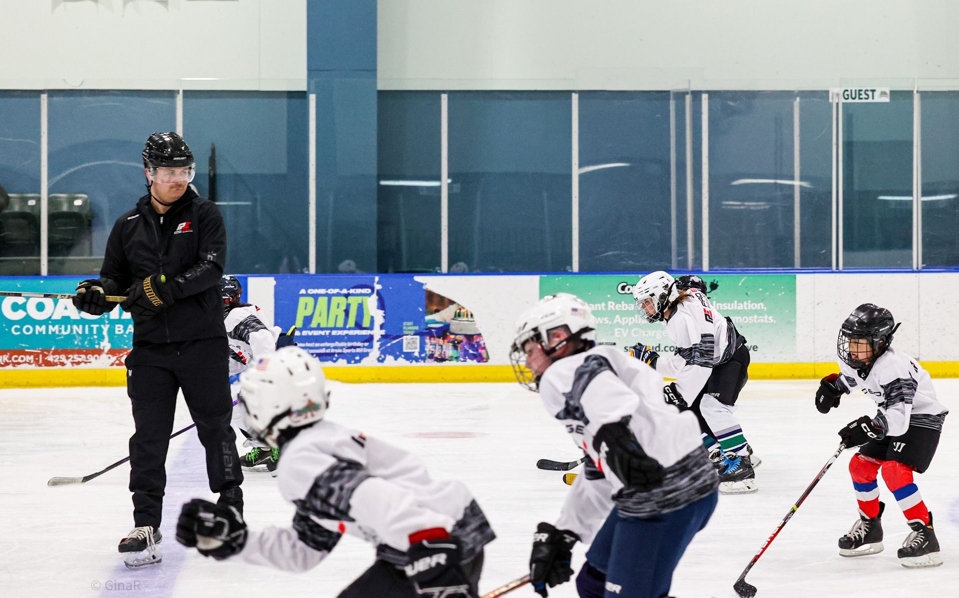 Youth hockey practice on an indoor rink, coach watching skaters in white jerseys and black helmets