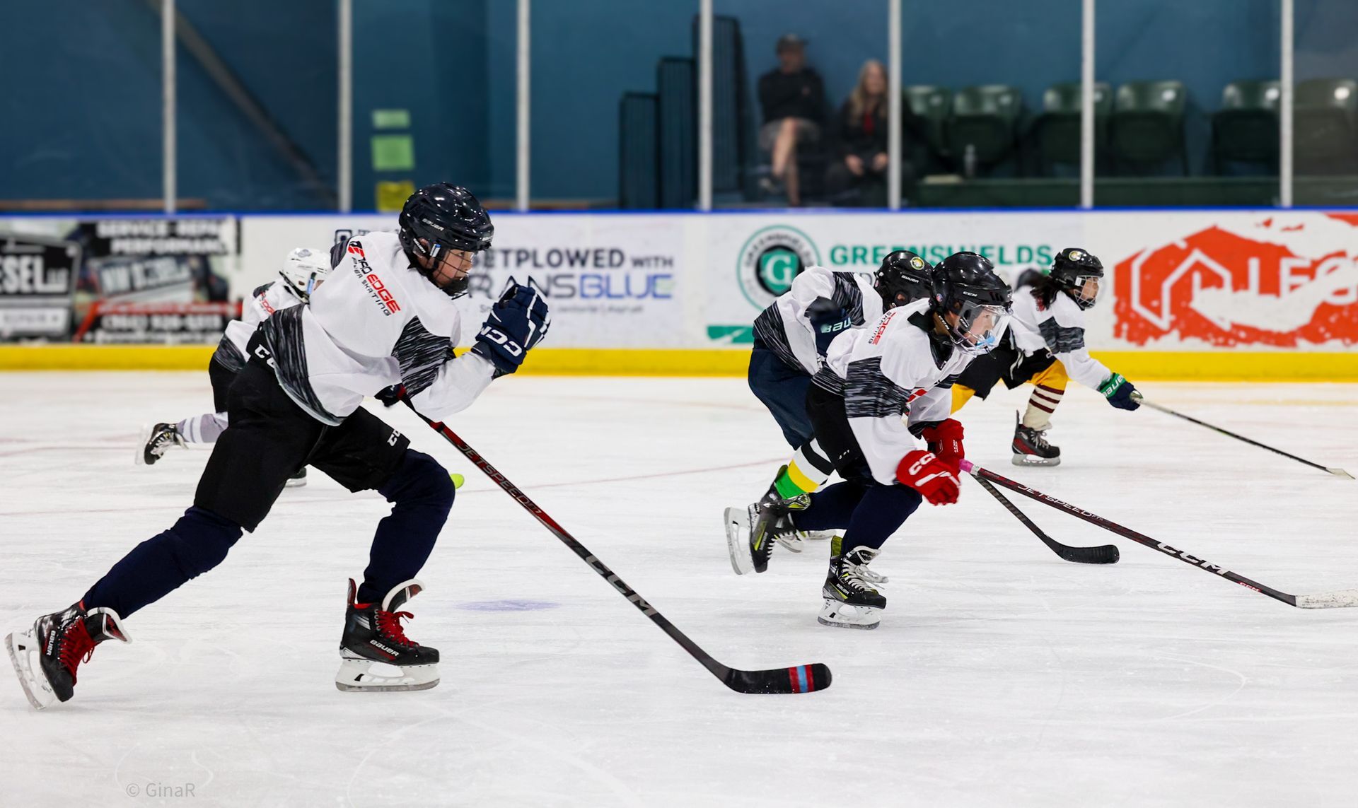 Ice hockey players in white and dark jerseys sprint after the puck on an indoor rink.