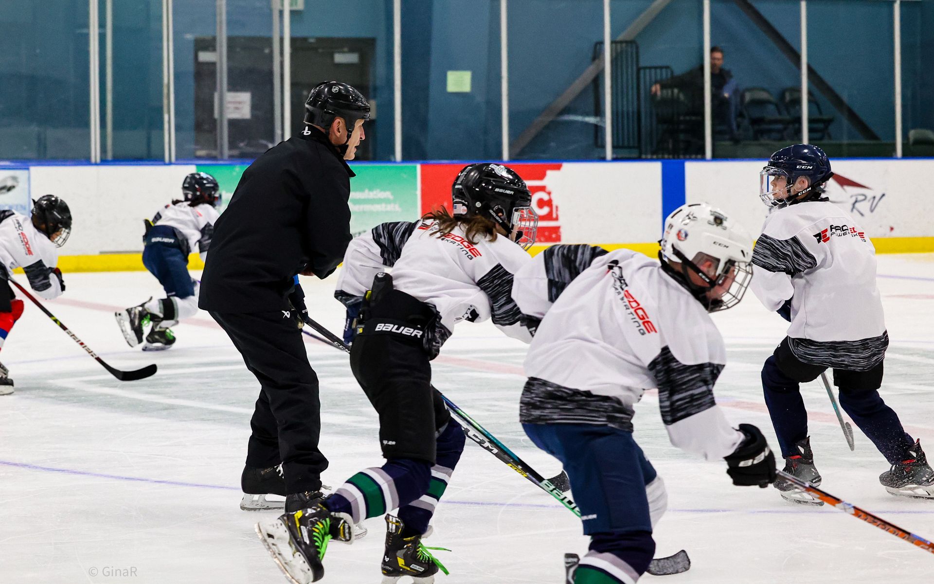 Youth hockey practice on an ice rink, with players in white jerseys and a coach in black skating alongside them.