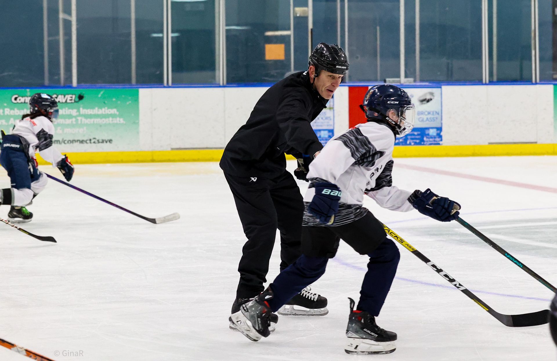 Two hockey players in white and black skate on an indoor rink while a coach watches nearby.