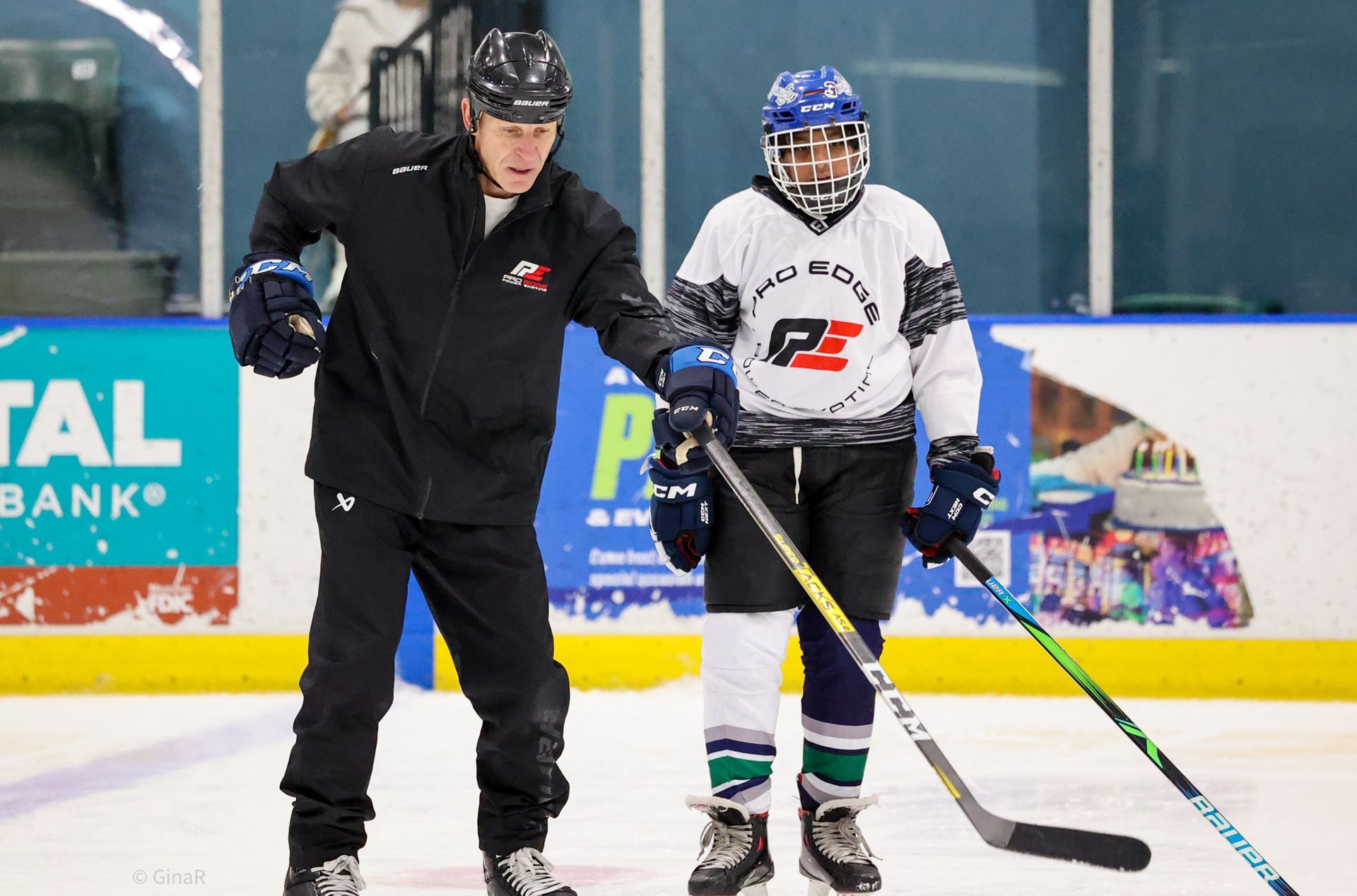 Two hockey players on ice, one in black coaching and one in white jersey holding a stick.