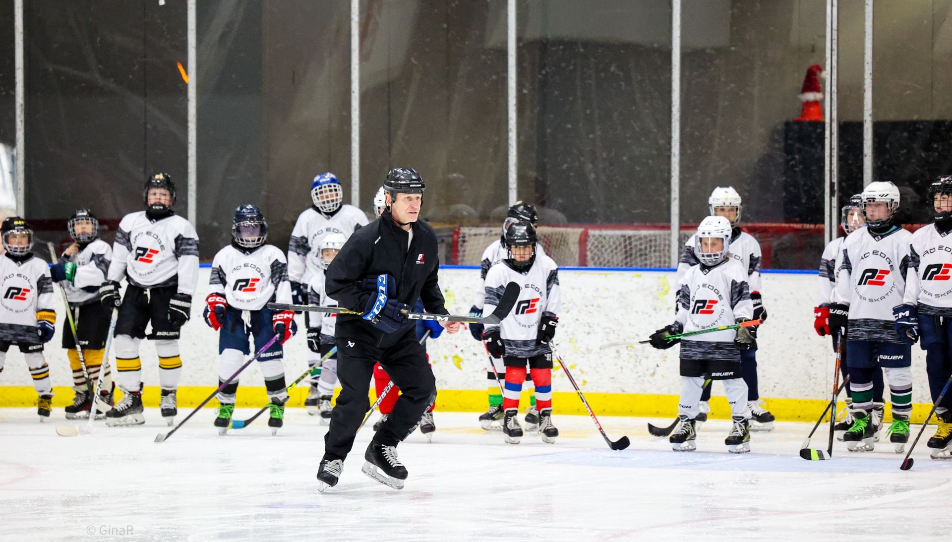 Youth hockey players in white jerseys line up on the ice while a coach skates in front of them.
