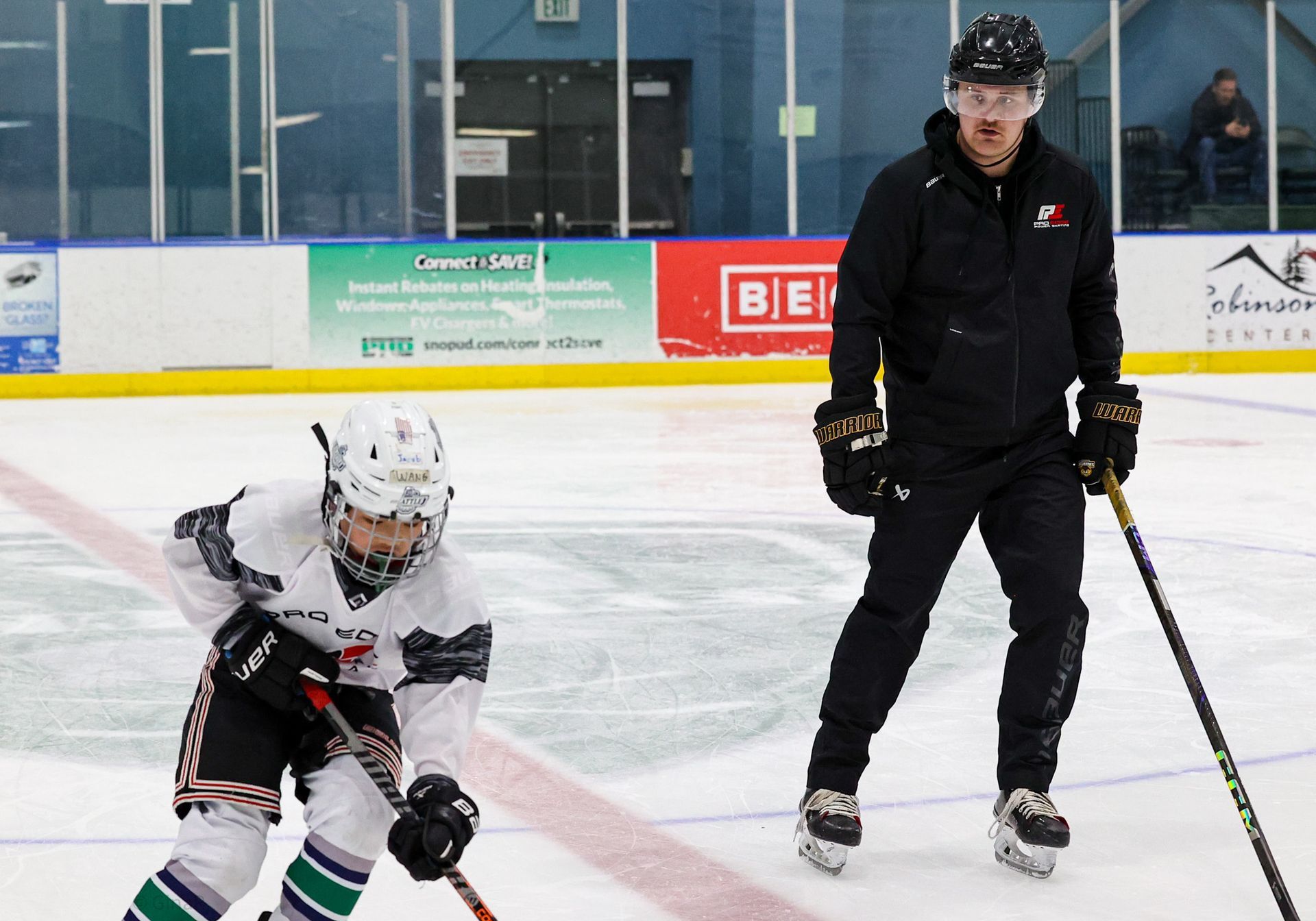 Young hockey player crouches on ice beside coach in black gear at indoor rink