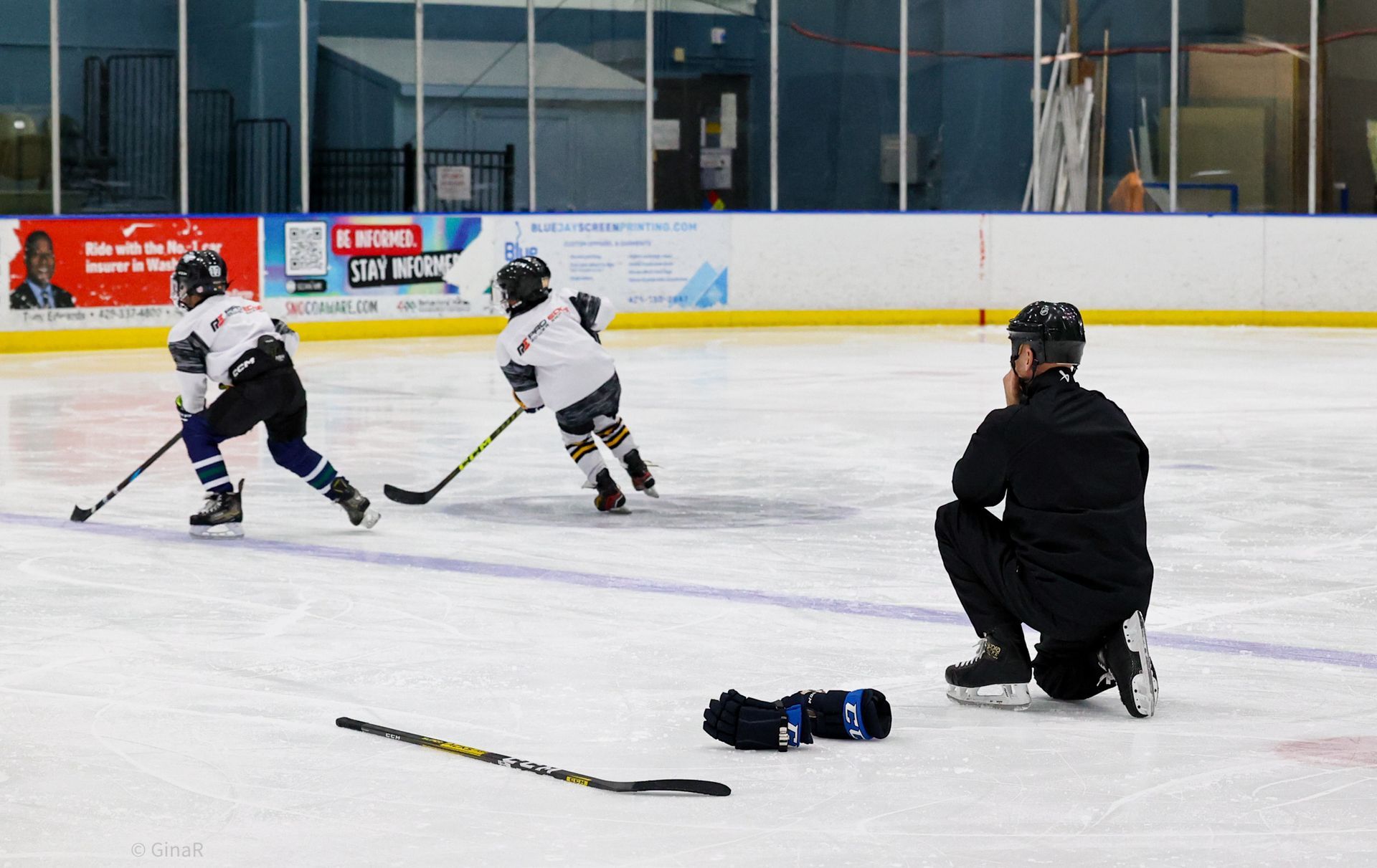 Two hockey players in white practice on an indoor rink while a coach kneels nearby.
