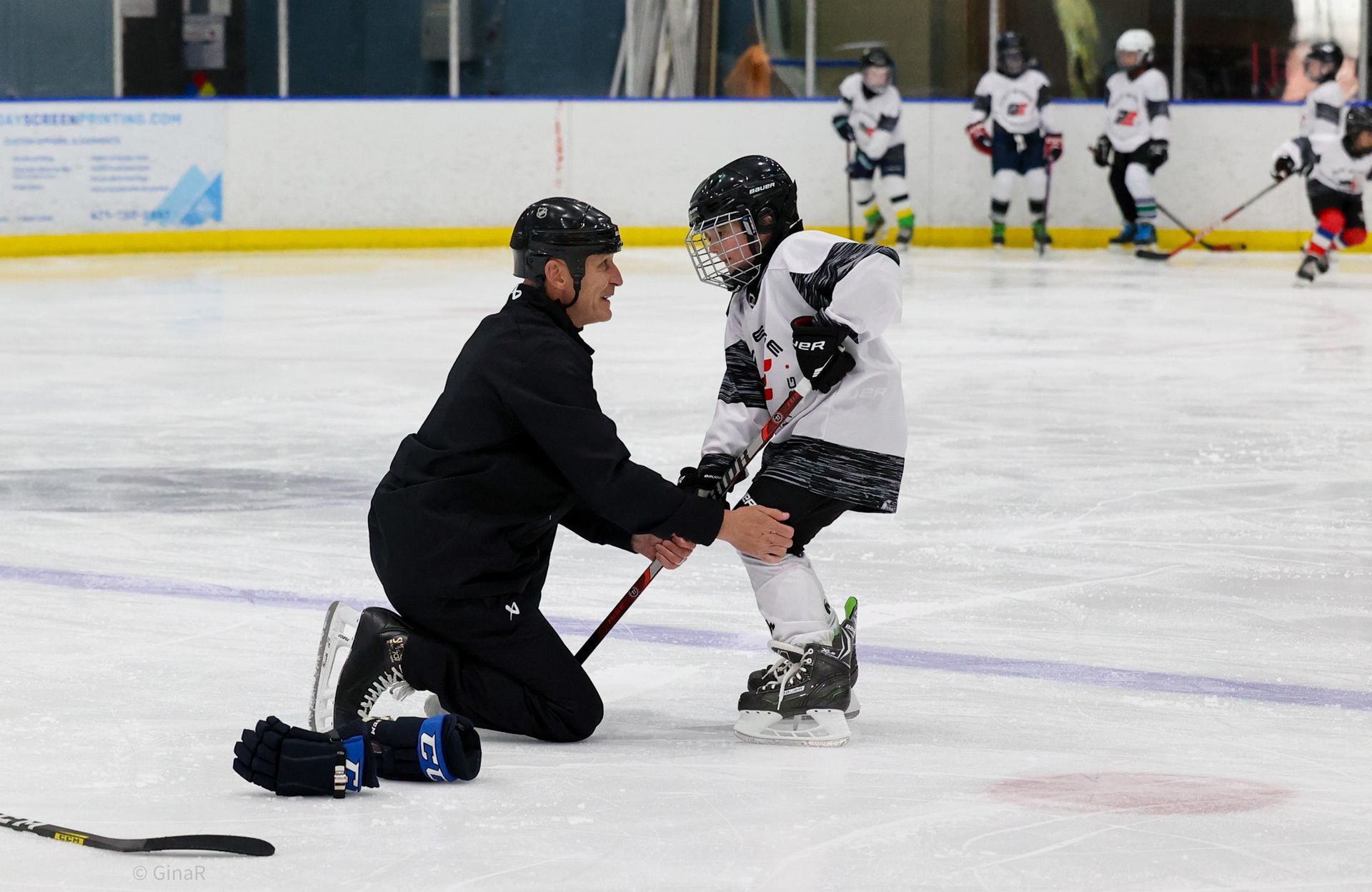 Coach kneels on the ice helping a young hockey player balance during practice; teammates watch nearby.