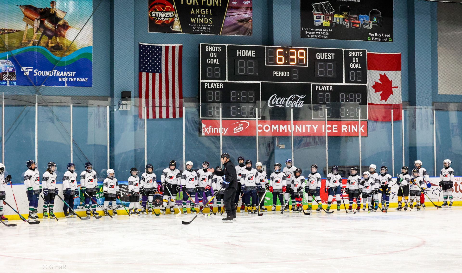 Ice hockey teams lined up on the bench at an indoor rink beneath the scoreboard and flags.