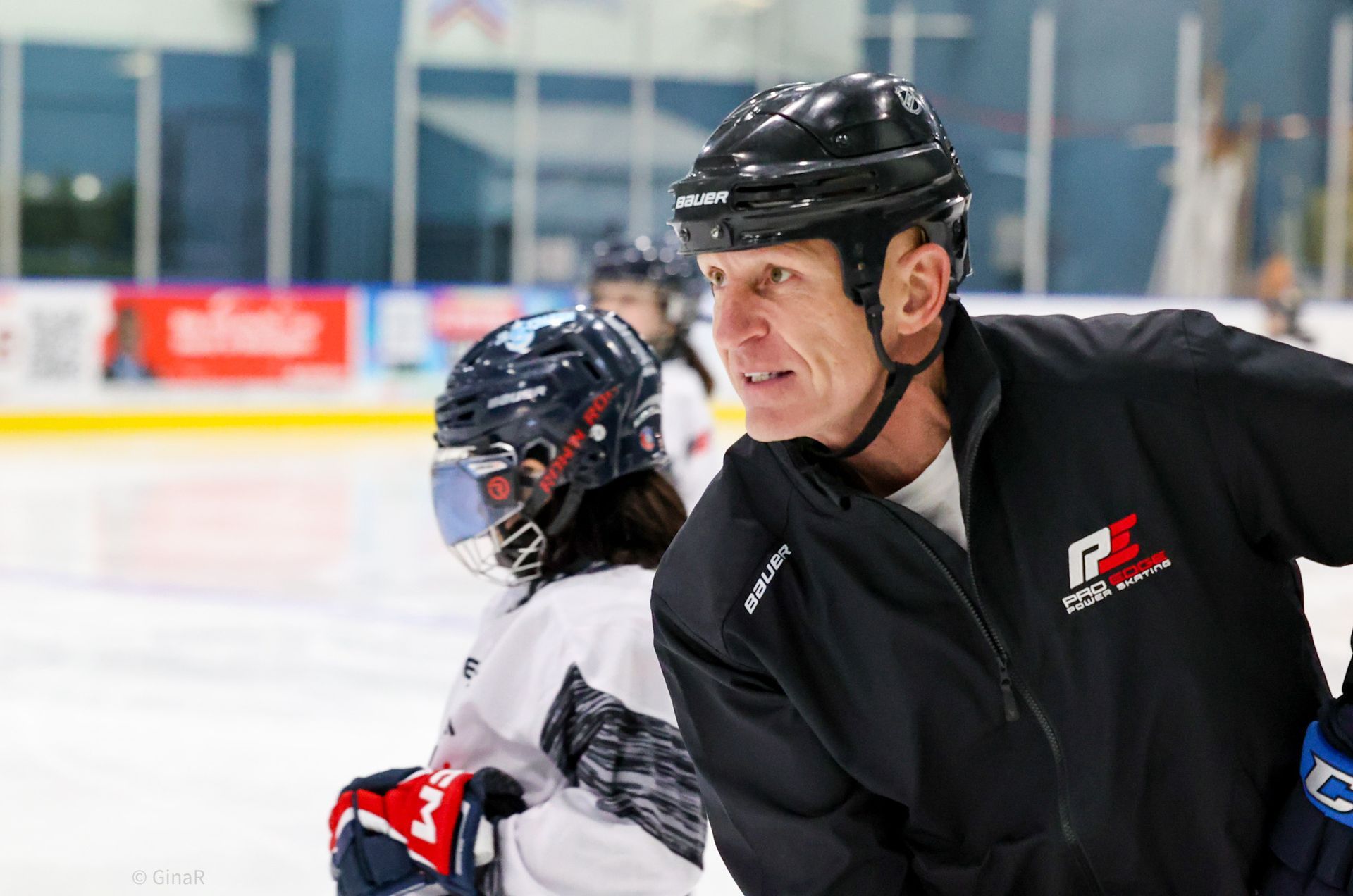 Two ice hockey players in helmets on the rink, one in black leaning forward in the foreground.