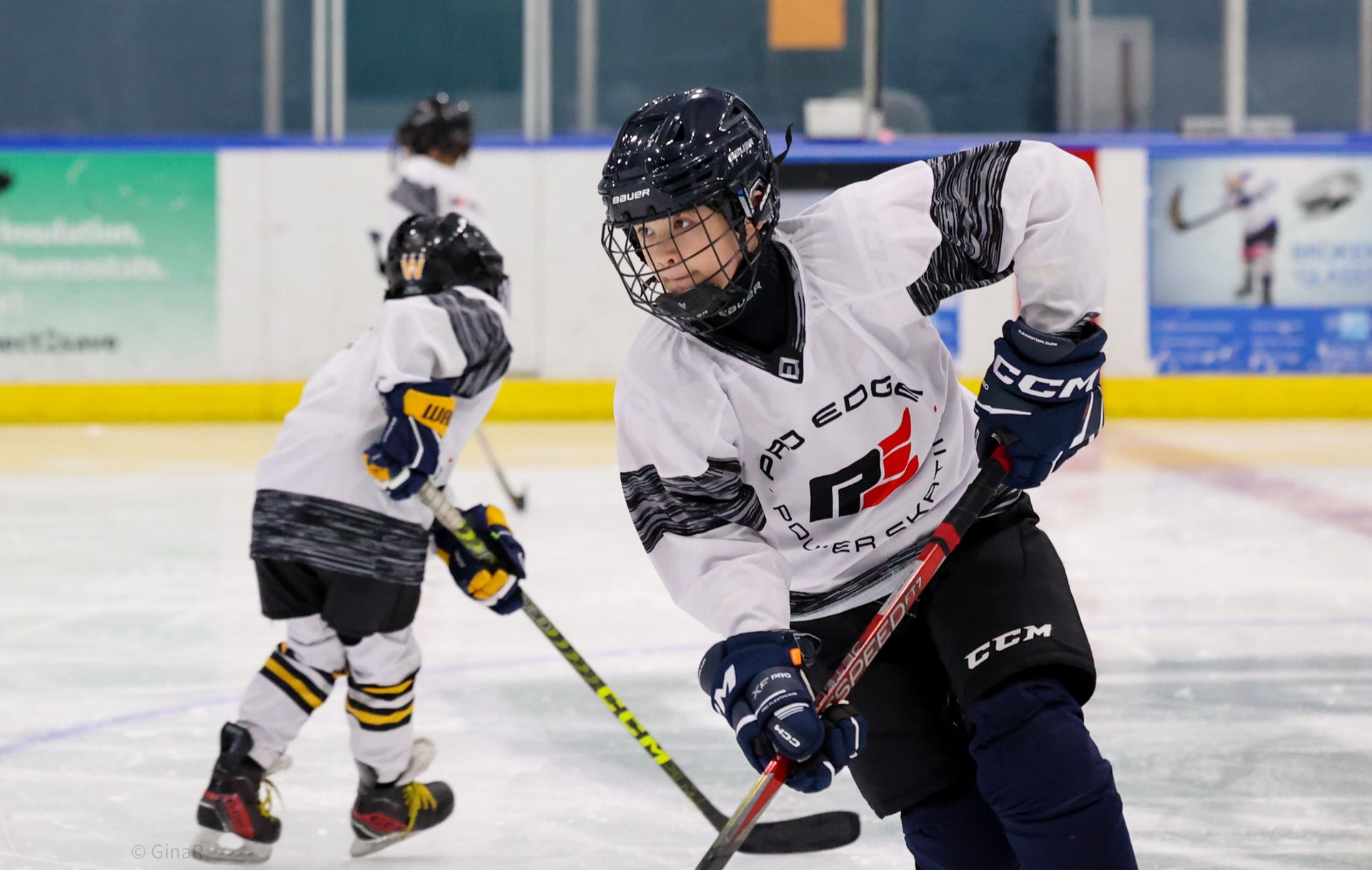 Youth ice hockey players in white jerseys skating on an indoor rink, one in the foreground with a stick.