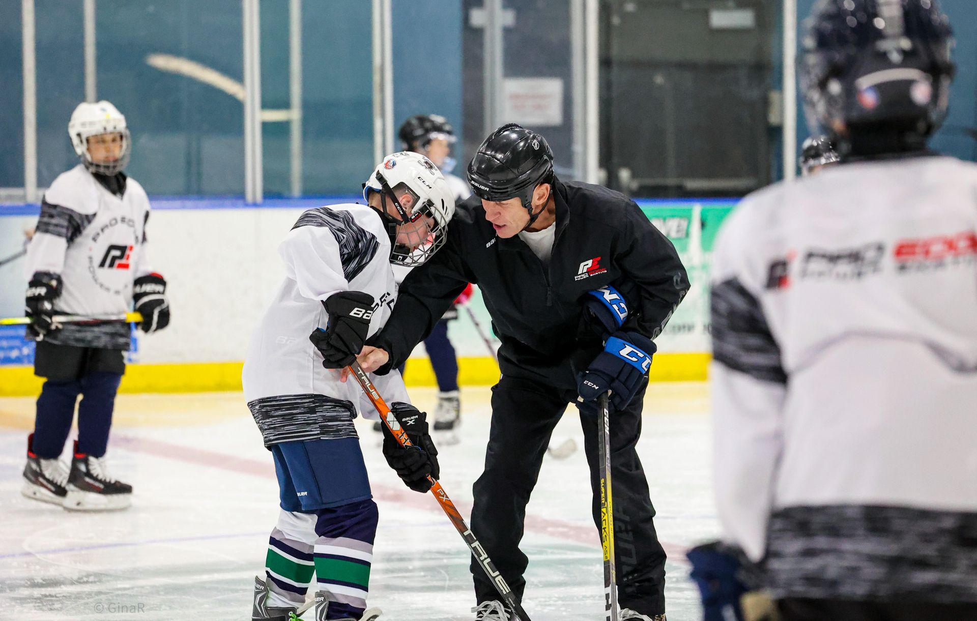 Ice hockey players practice faceoff on an indoor rink, with a coach in black assisting.