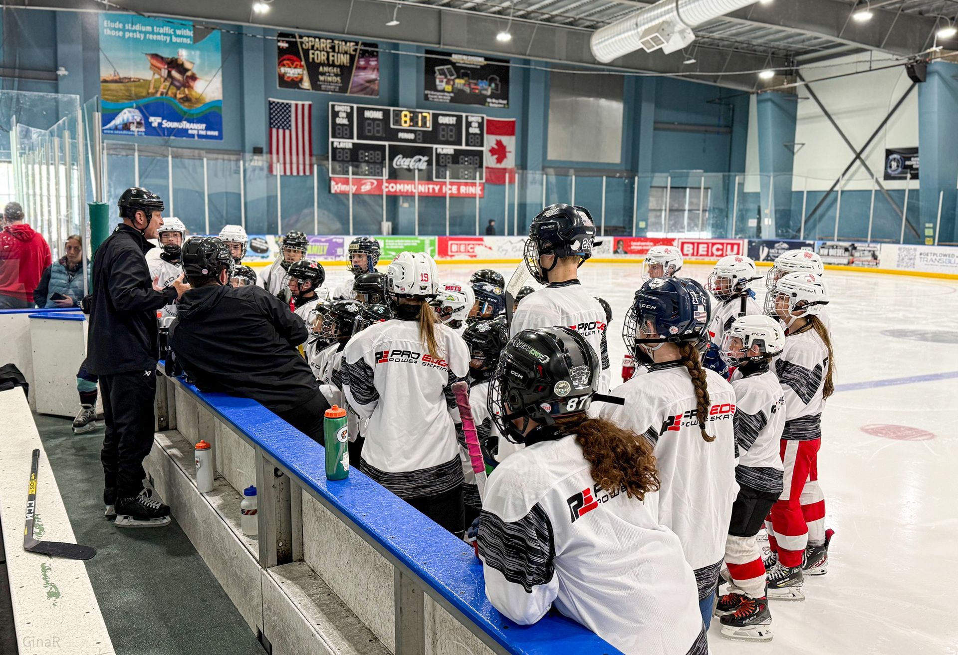 Youth hockey team on an indoor rink bench, players in white and black gear watching the game.