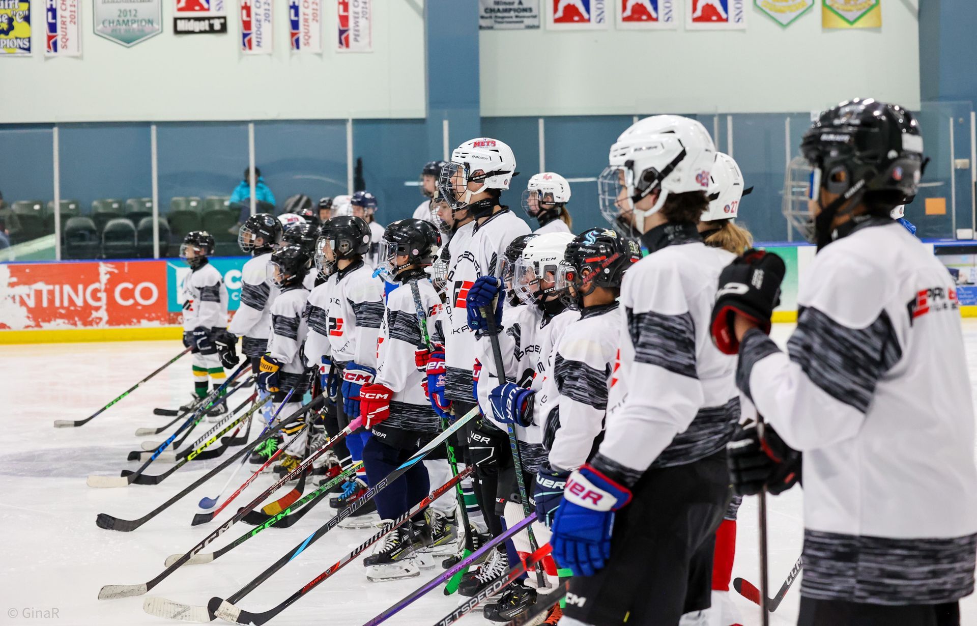 Youth hockey players in white jerseys lined up on an indoor rink, holding sticks and facing left
