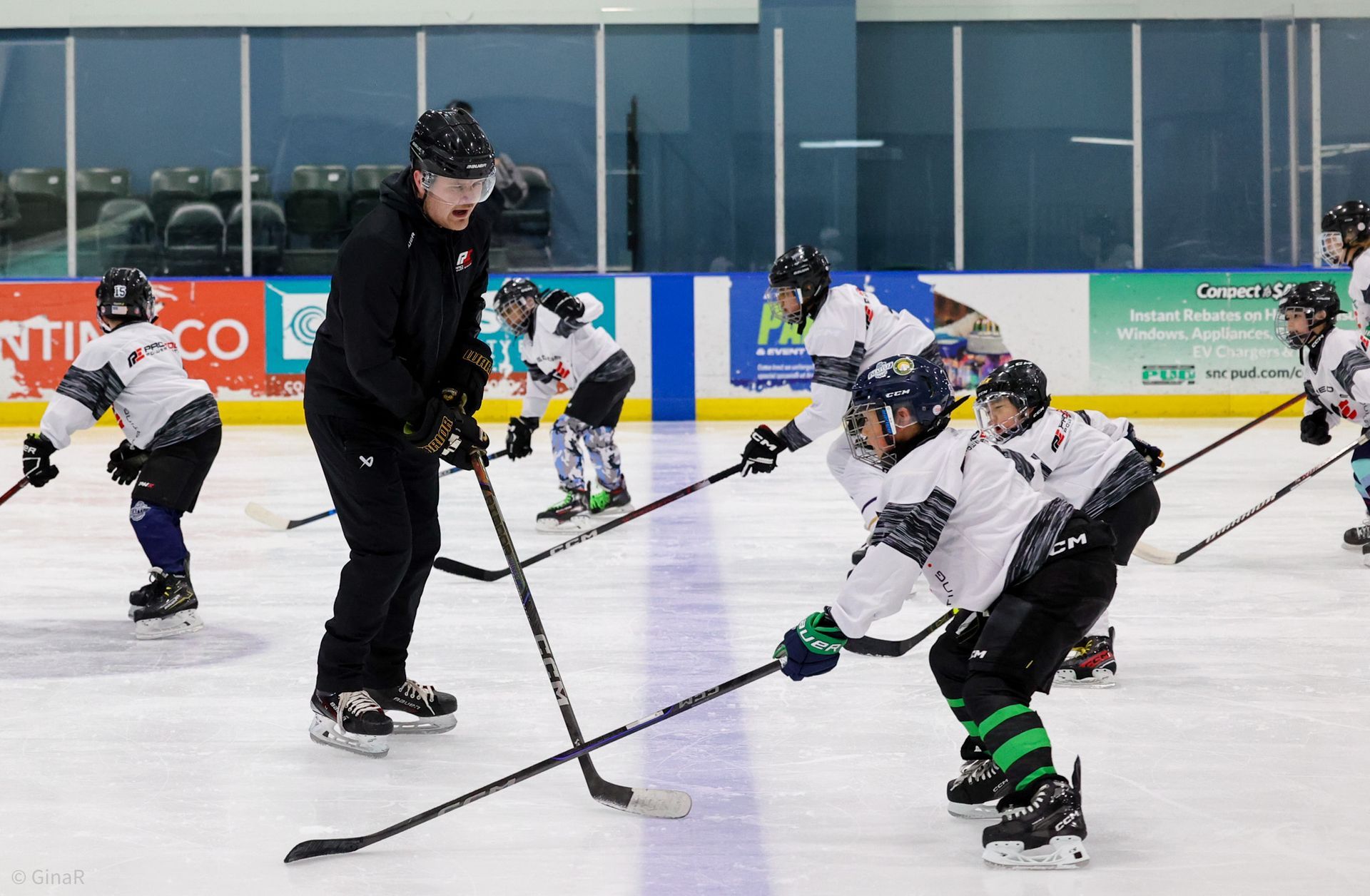Youth hockey coach instructs skaters on an indoor rink during practice, with players crouched in gear.