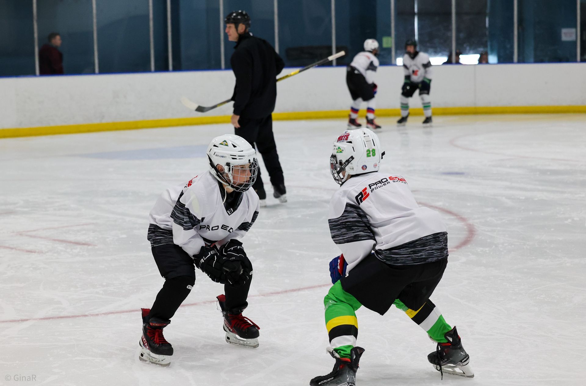 Two hockey players in white jerseys face off on an indoor ice rink, with a coach and teammates in the background