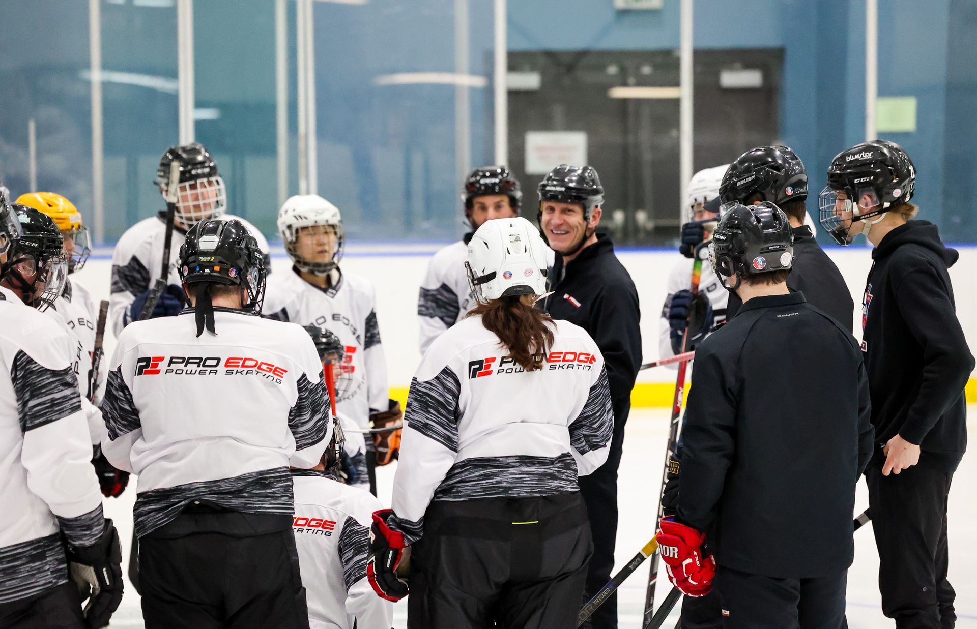 Women in hockey gear huddle on an ice rink, listening to a coach.