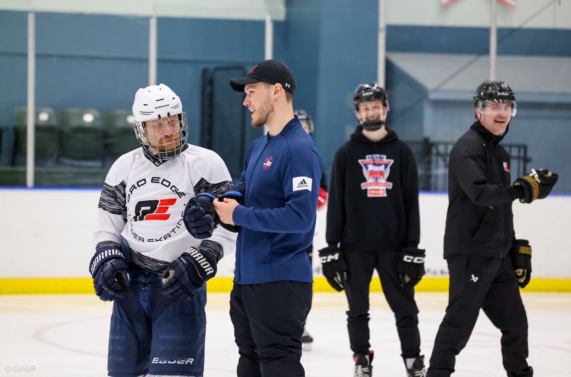 Four hockey players on an ice rink, one in a white jersey and three in dark gear, standing and talking