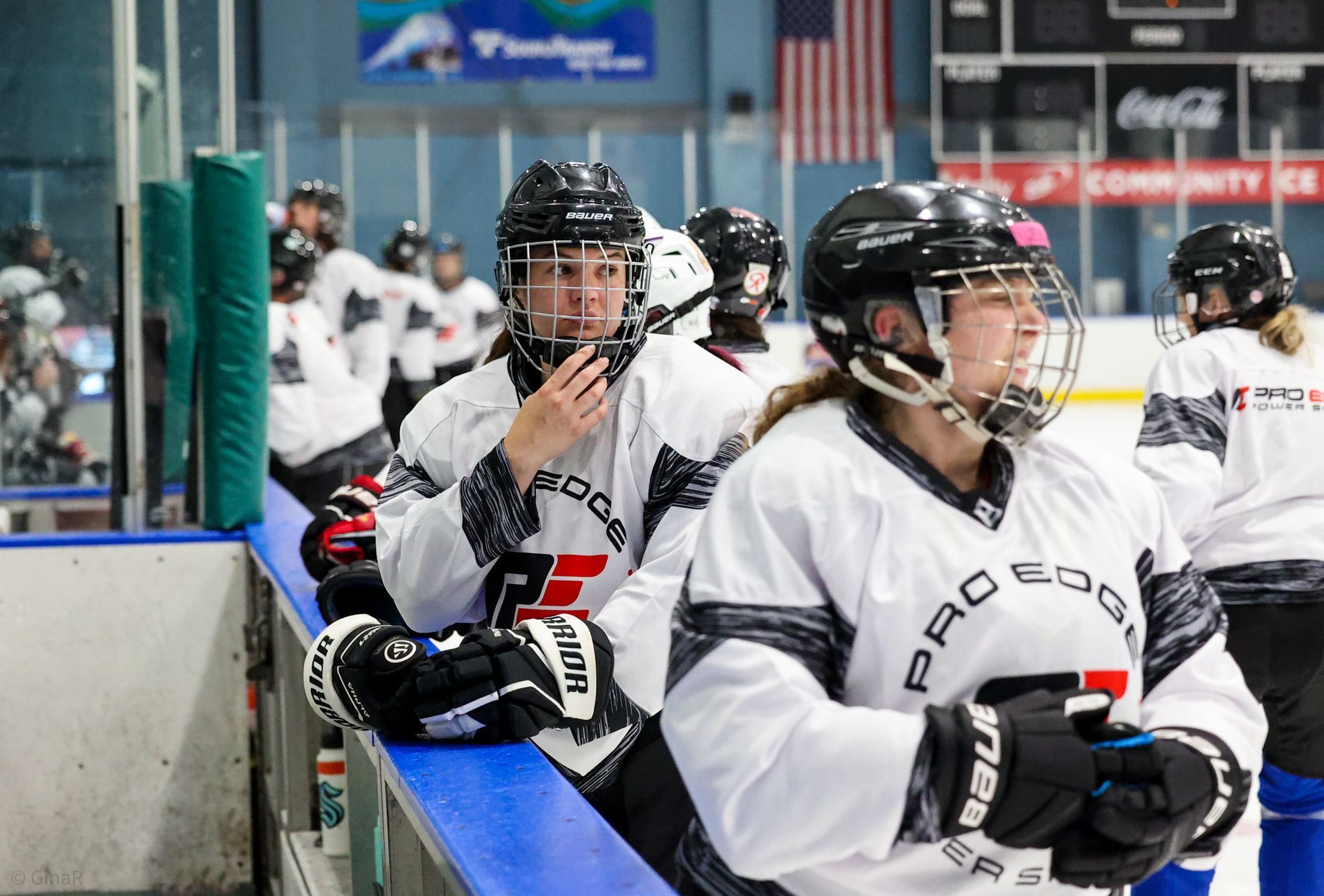 Hockey players in white jerseys sit on the bench during a game, one looking toward the ice.