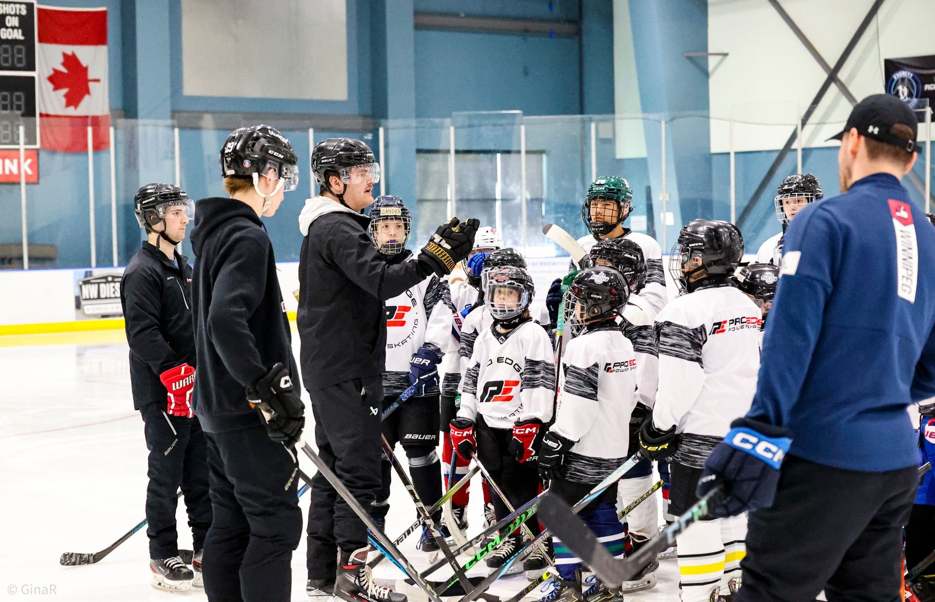 Youth hockey team huddles on an indoor rink while a coach in blue speaks to them near the boards.