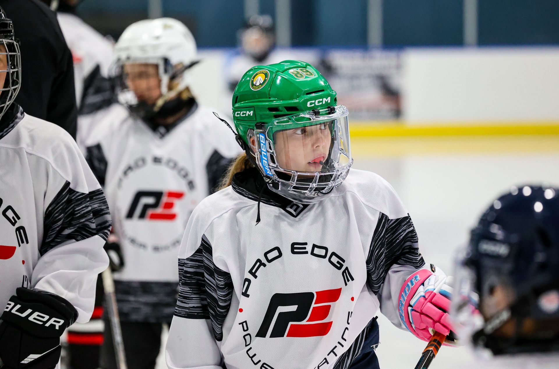 Youth ice hockey players in white jerseys on the rink, one in a green helmet and pink gloves