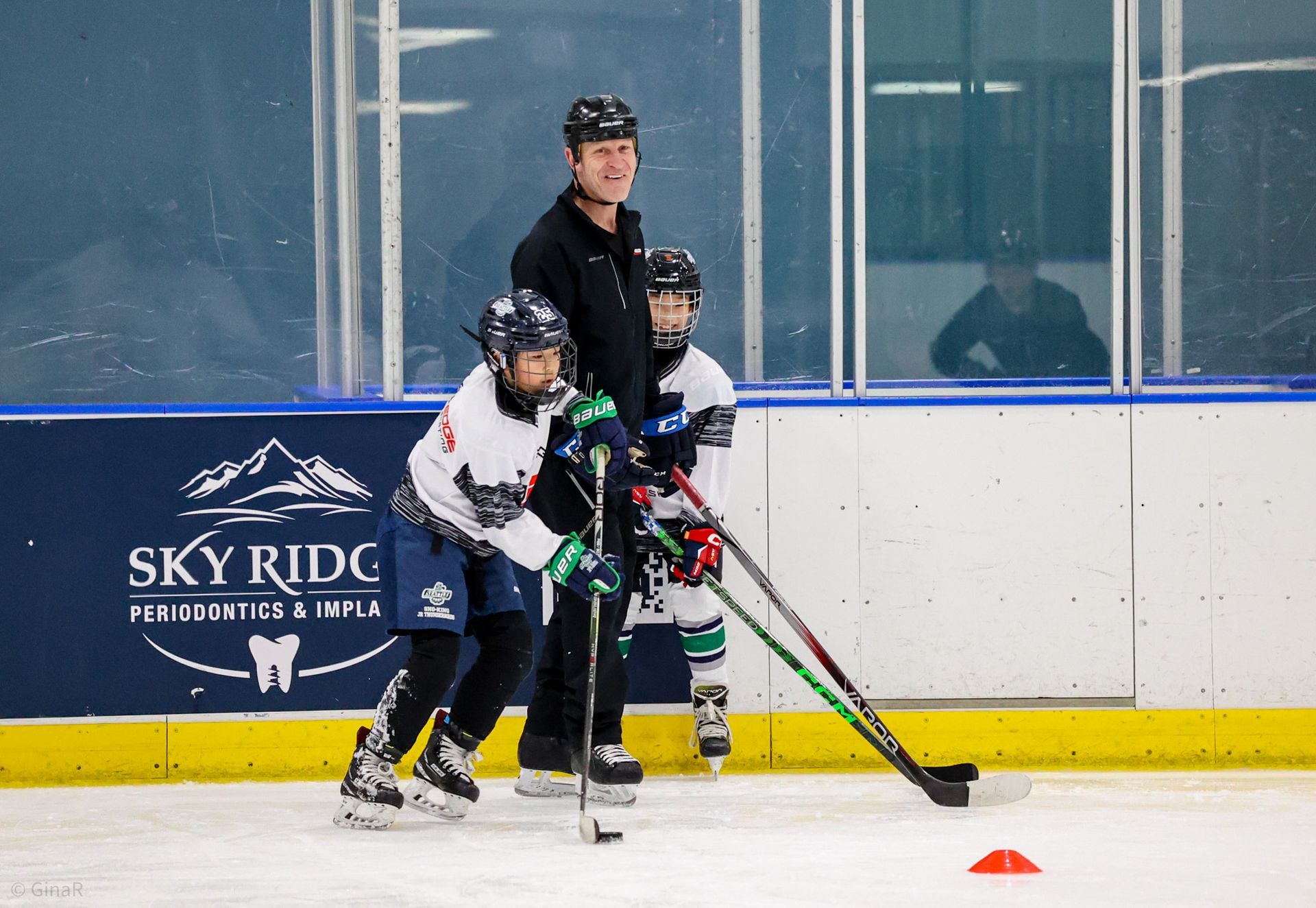 Three hockey players practice on ice near orange cones, with one coach behind them.