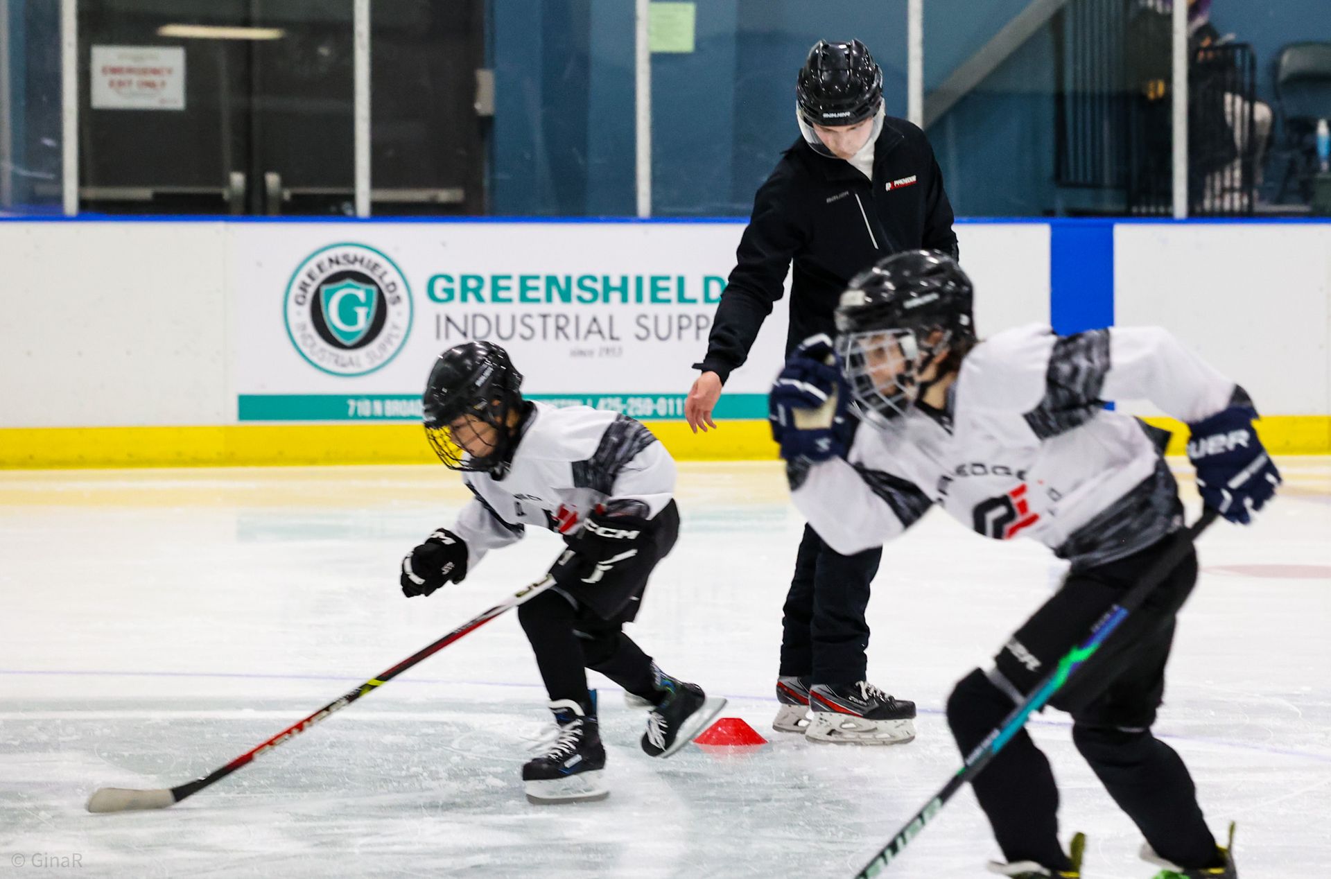 Youth hockey players in black and white practice on an ice rink with a coach behind them.