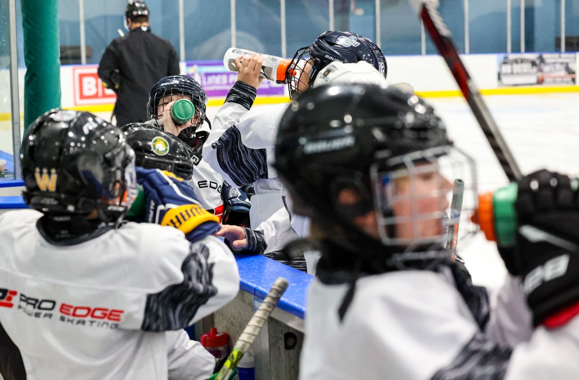 Youth hockey players in helmets crowd the rink, one raising a gloved hand near the boards.