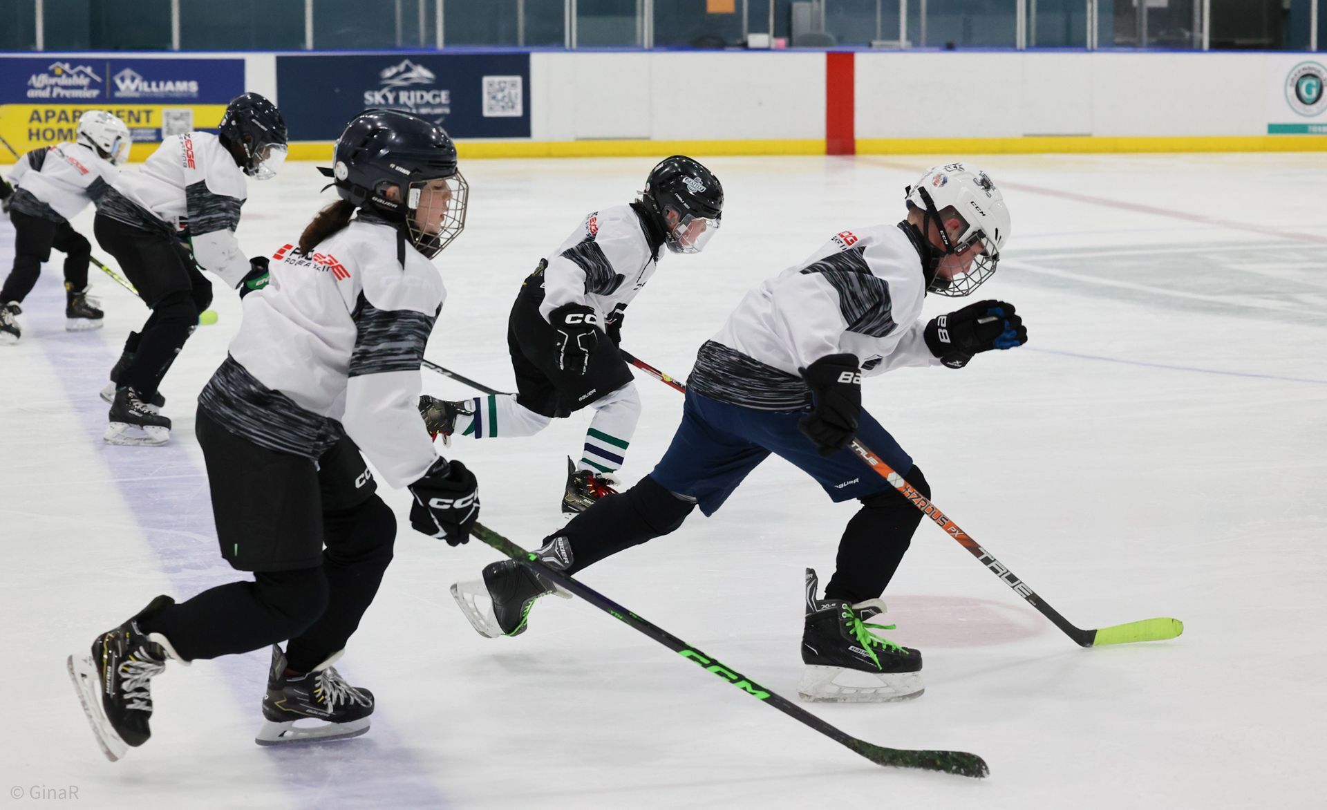Hockey players in white jerseys skating fast on an indoor ice rink, chasing the puck