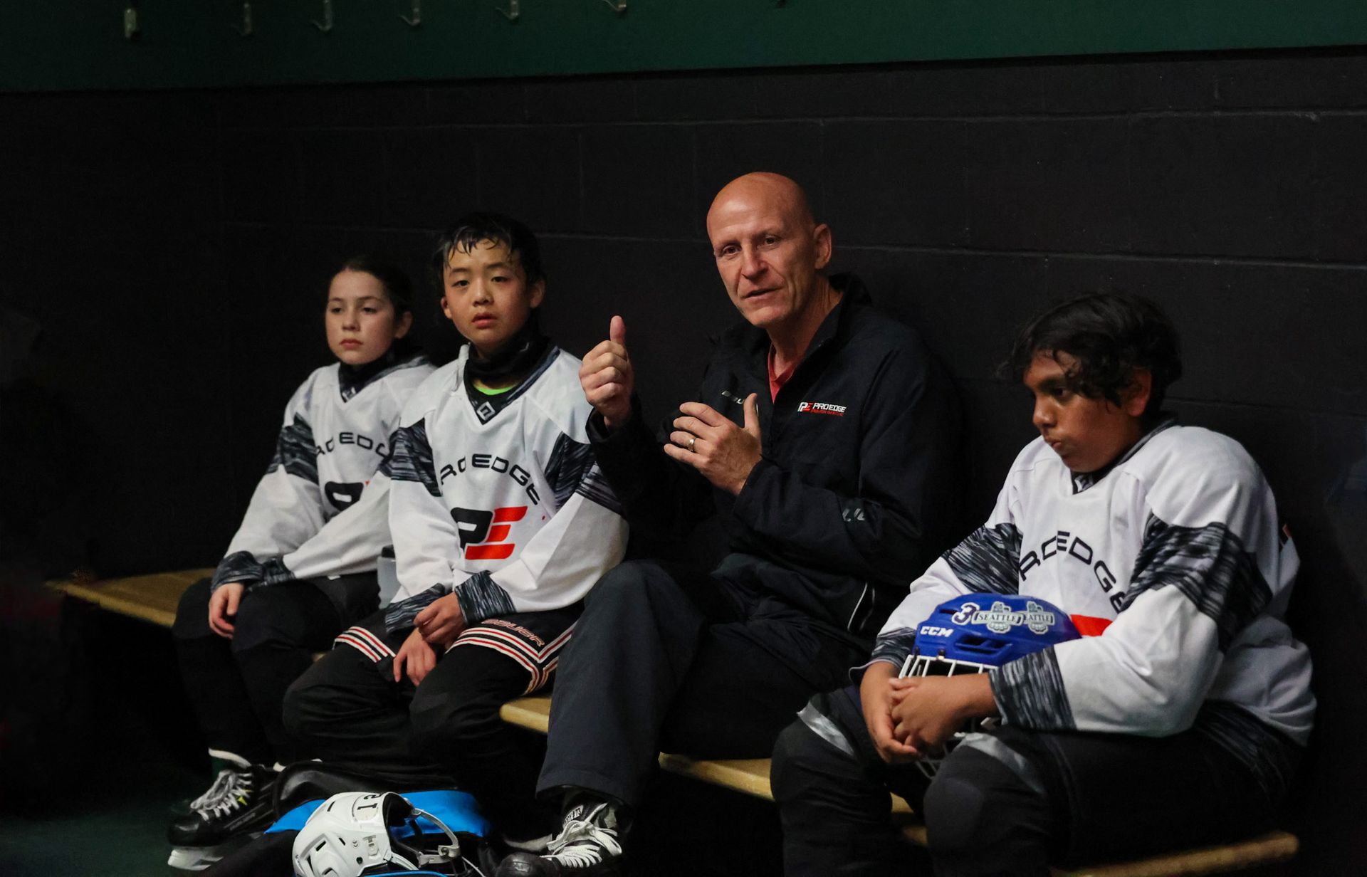Four people in white martial arts uniforms sitting on a bench in a dark locker room, one giving thumbs up