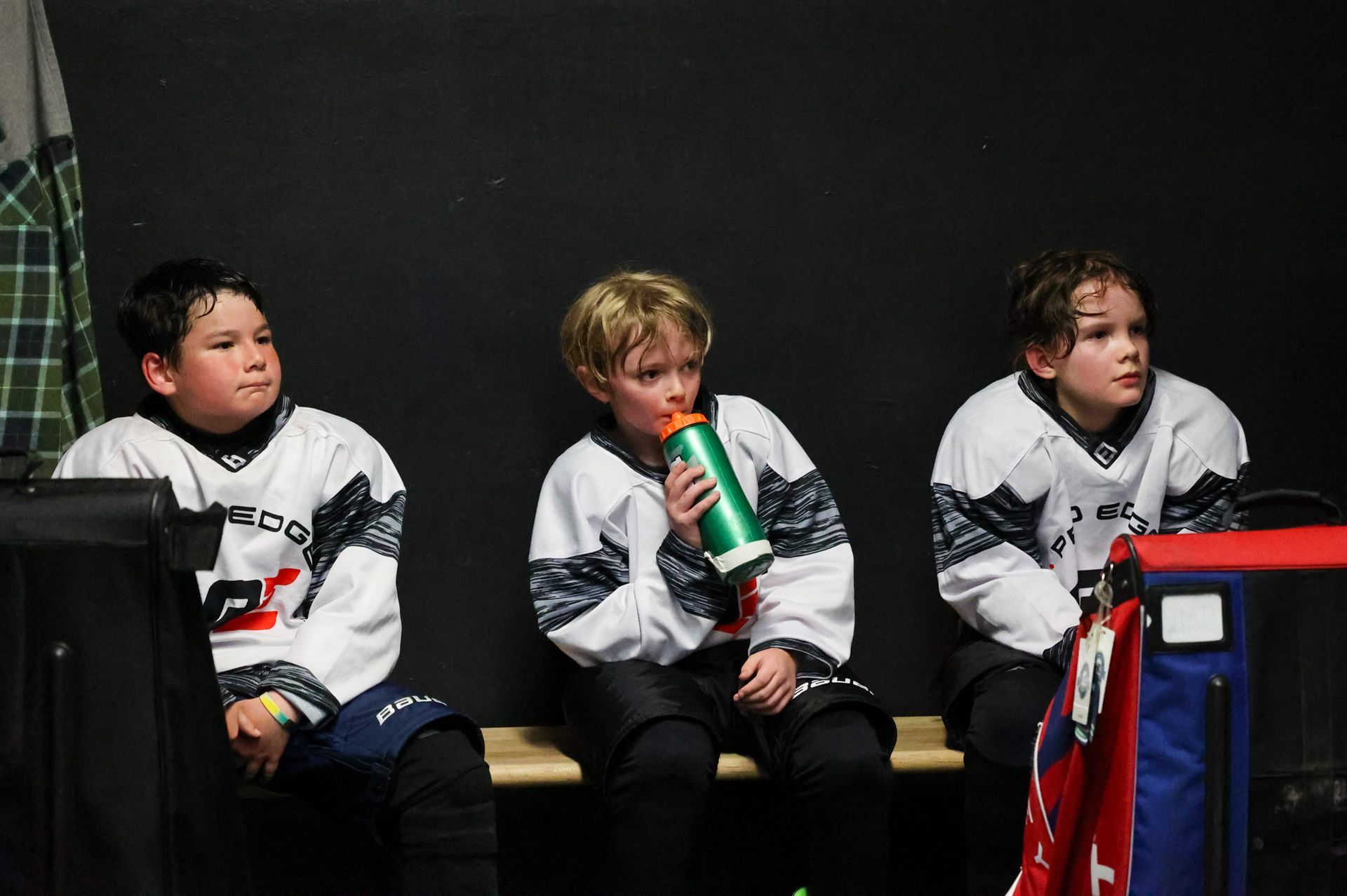 Three youth hockey players sit on a bench, one drinking from a green water bottle.